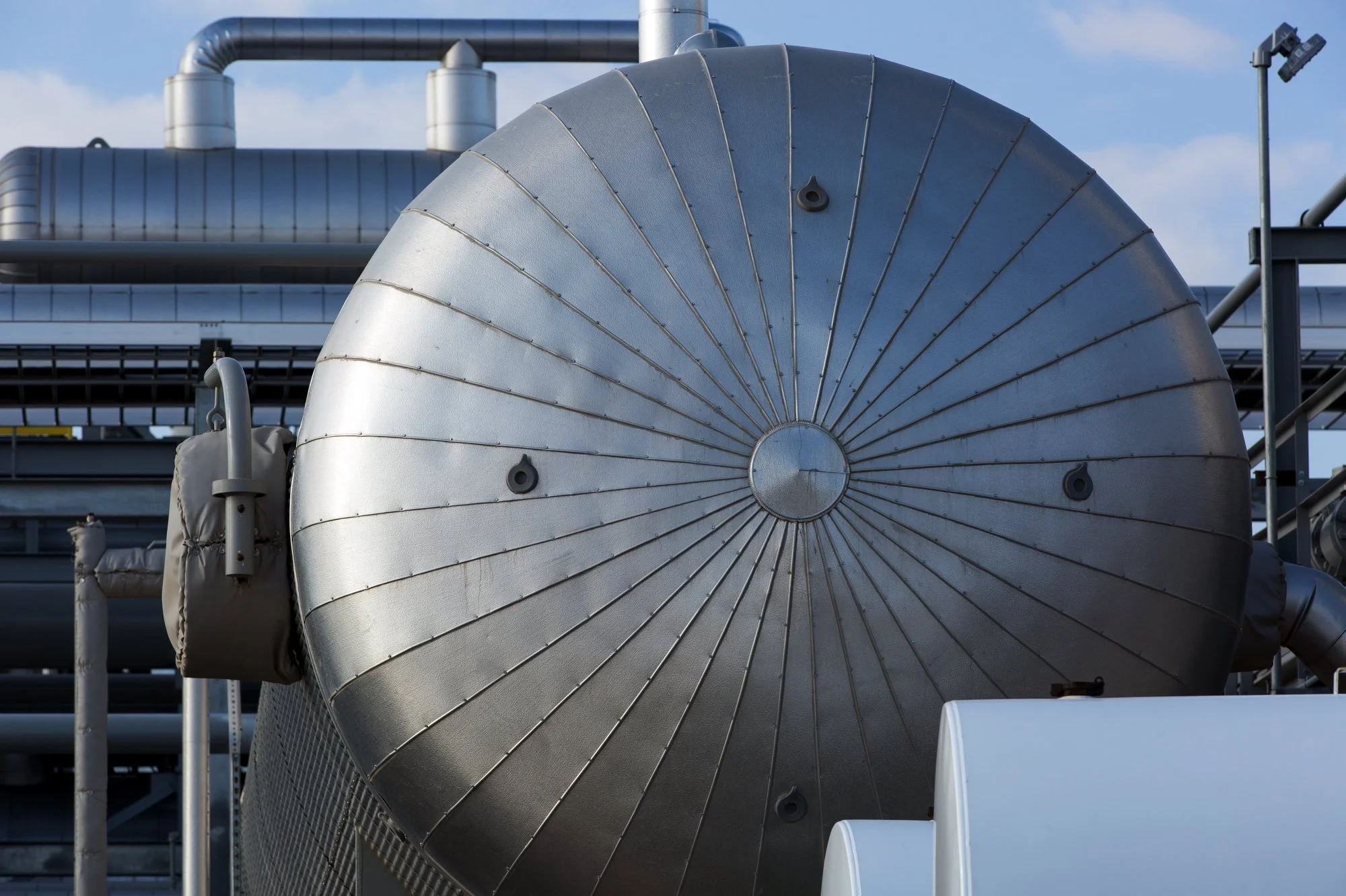 Close-up of a large, metallic industrial tank or vessel on an industrial site, surrounded by pipes and equipment under a clear blue sky.