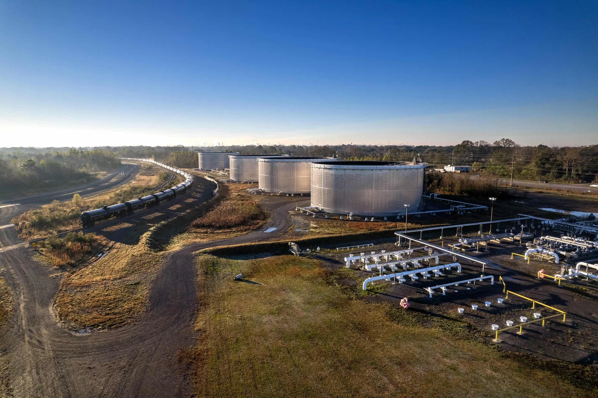 An aerial view of large industrial storage tanks and pipelines at an industrial facility next to a railway track, with a clear blue sky overhead.