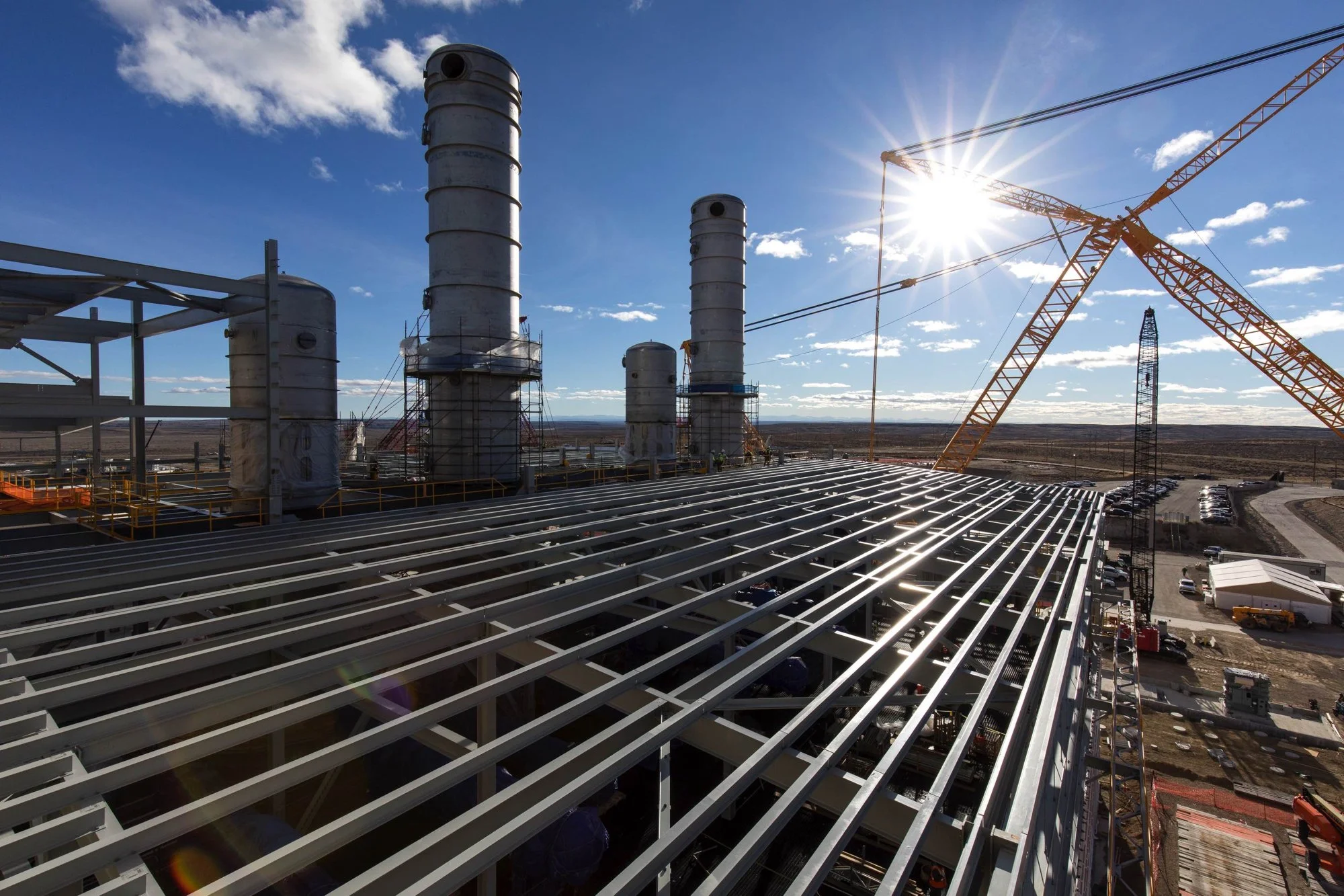 Construction site with industrial structures, tall pipes, a crane, and solar panels under a clear sky with the sun shining.