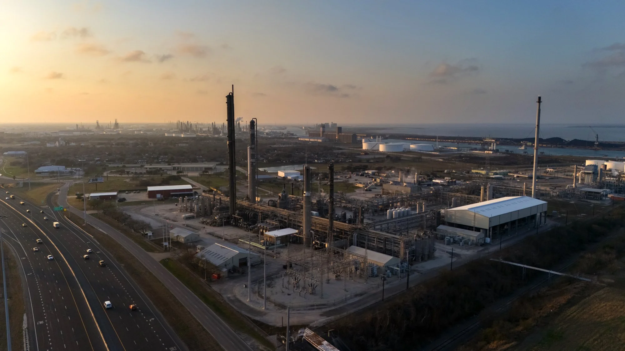 An industrial oil refinery facility near a highway, with multiple tall distillation columns, storage tanks, and buildings, set against a backdrop of open land, water, and a distant city skyline at sunset.