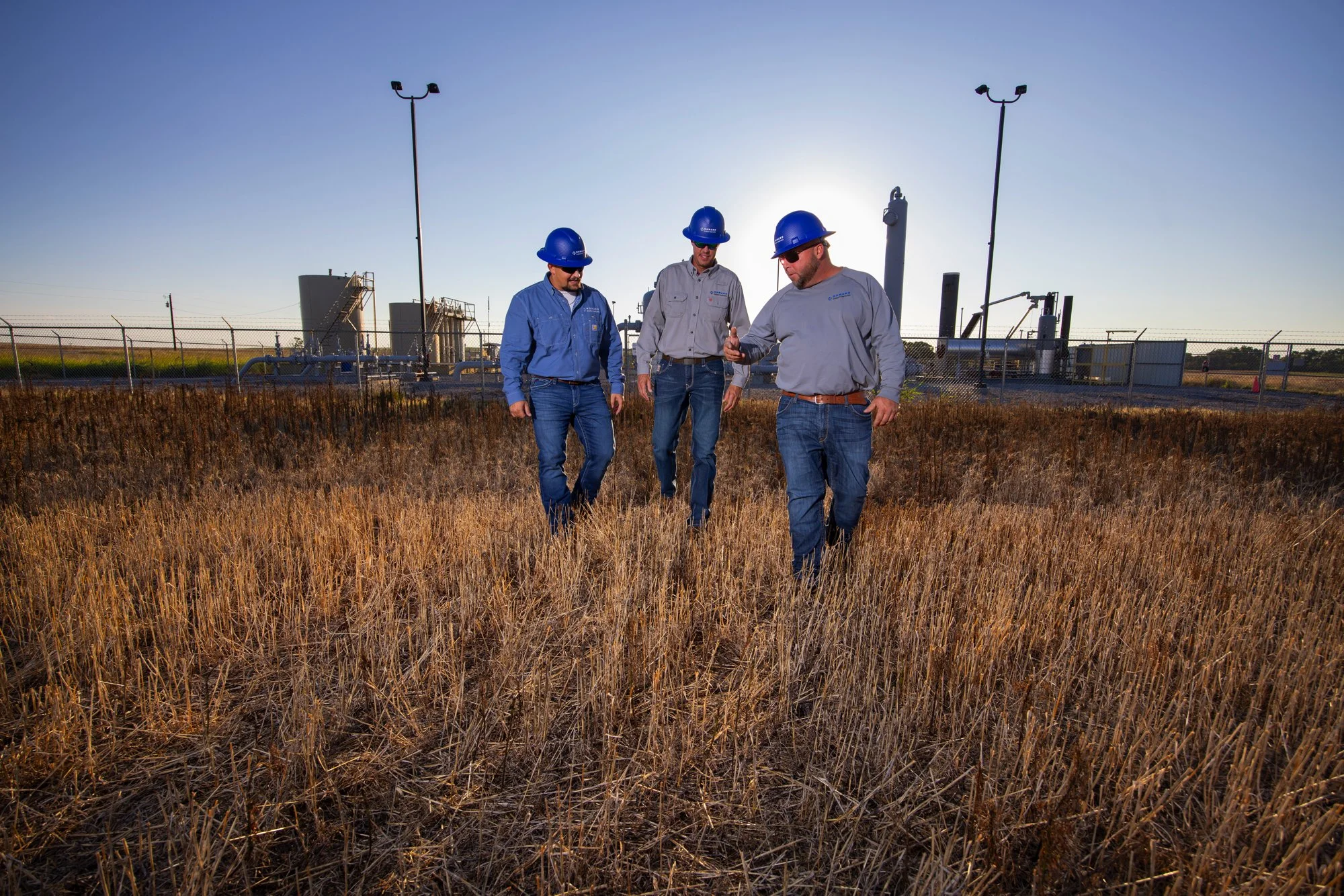 Three men in hard hats and jeans walking through a dry, grassy field with industrial equipment in the background.