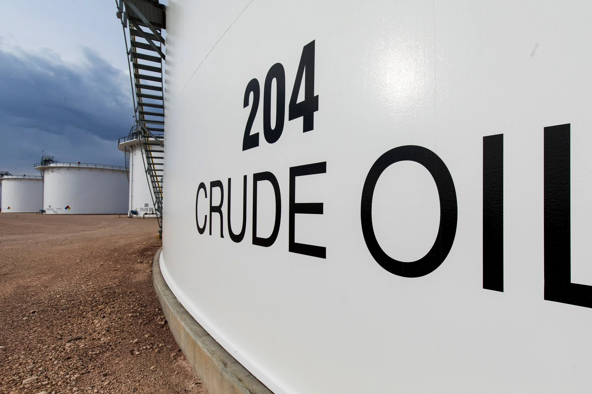 Oil storage tanks with the label '204 CRUDE OIL' in a refinery or storage facility, with a gravel ground and cloudy sky.