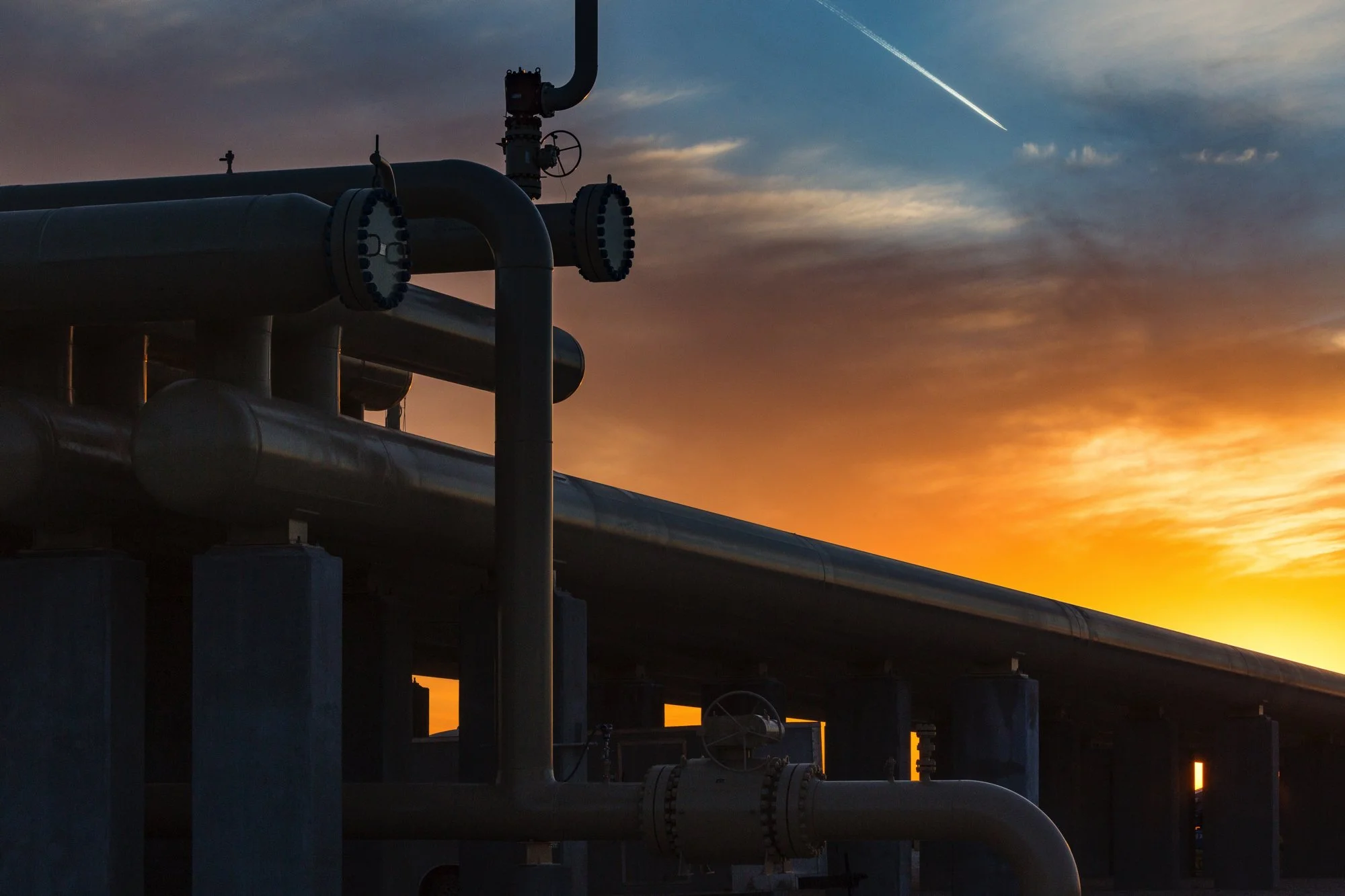 Industrial pipes and valves against a sunset sky with clouds and a visible contrail.