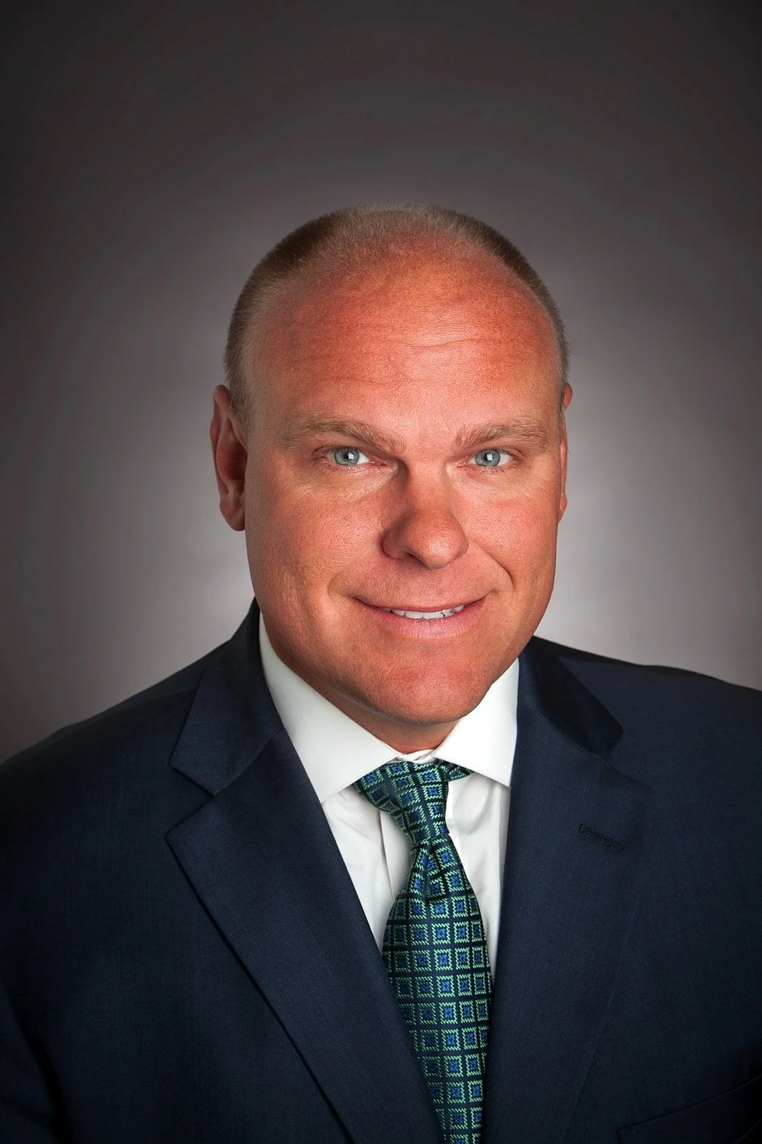 Professional headshot of a man in a suit, smiling, with a grey background.