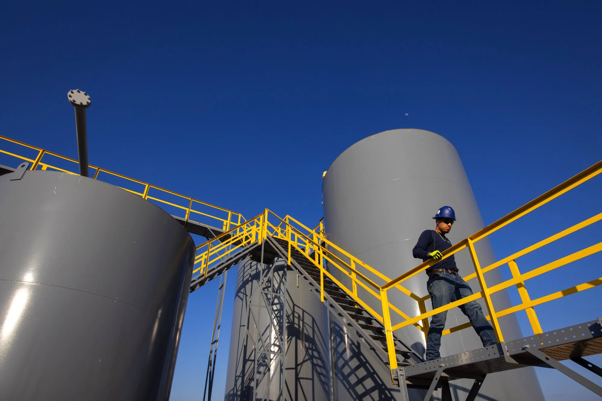 A worker wearing a helmet and gloves on a metal staircase with yellow railings, surrounded by large gray industrial tanks, against a clear blue sky.