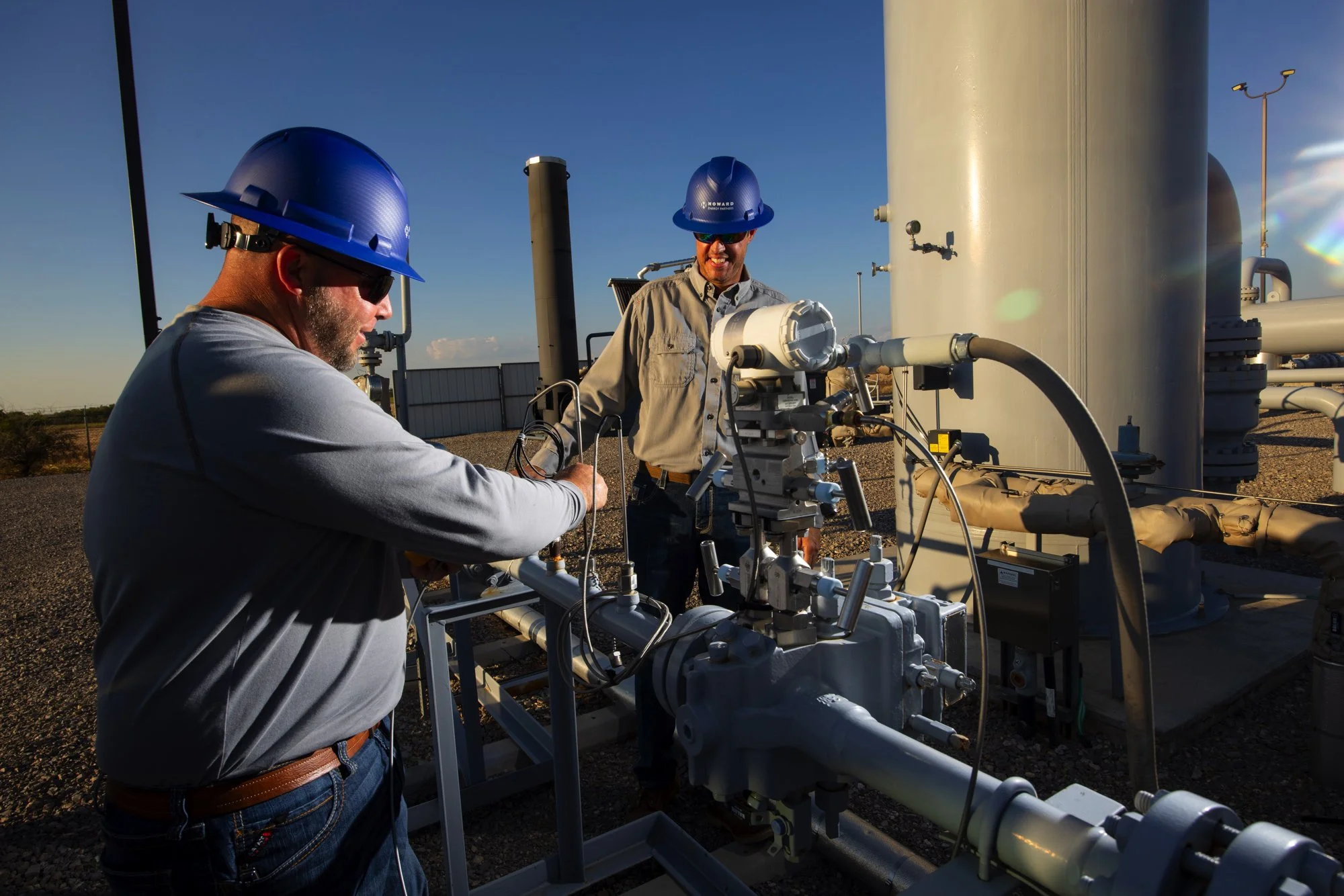 Two workers wearing blue hard hats and safety glasses working at an industrial site with pipelines and equipment under a clear sky.