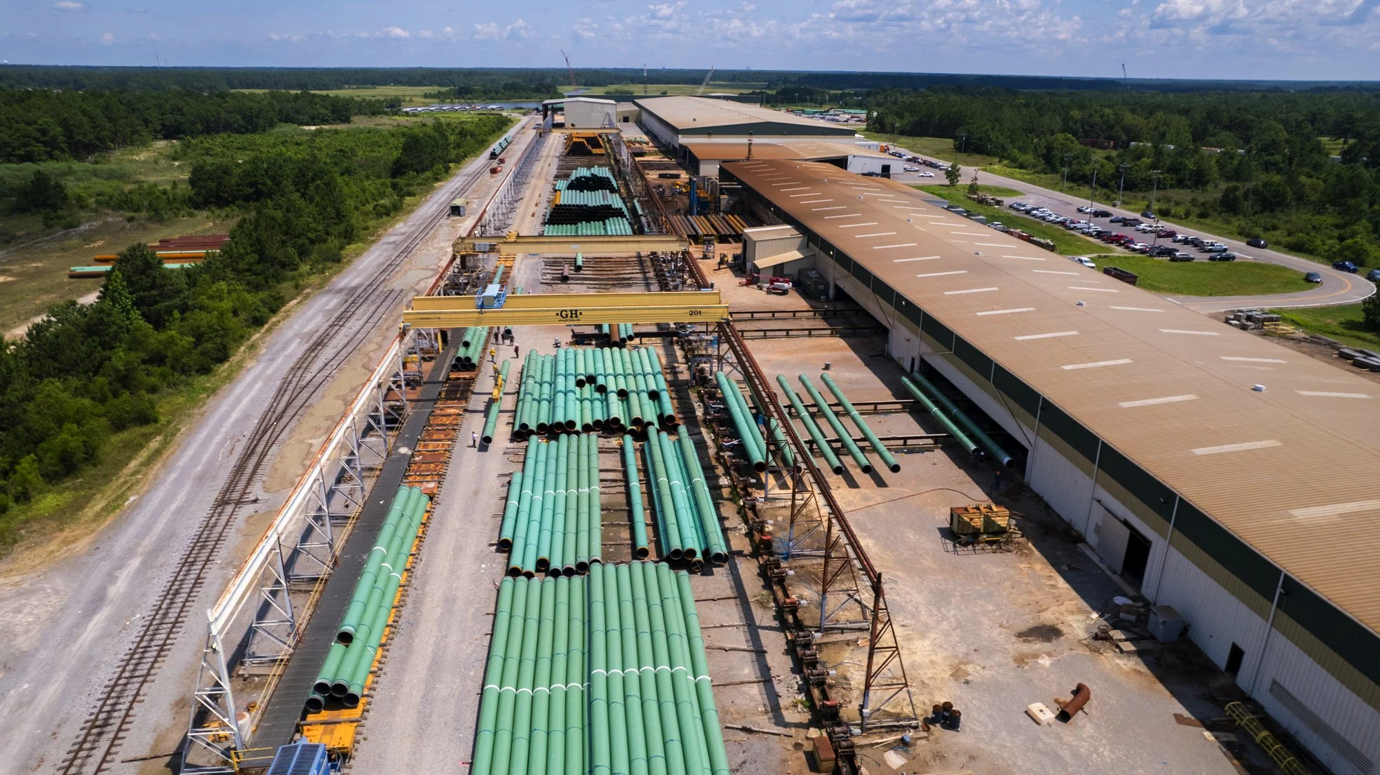 Aerial view of an industrial site with large green pipes, construction equipment, and a warehouse surrounded by trees and parking lot.