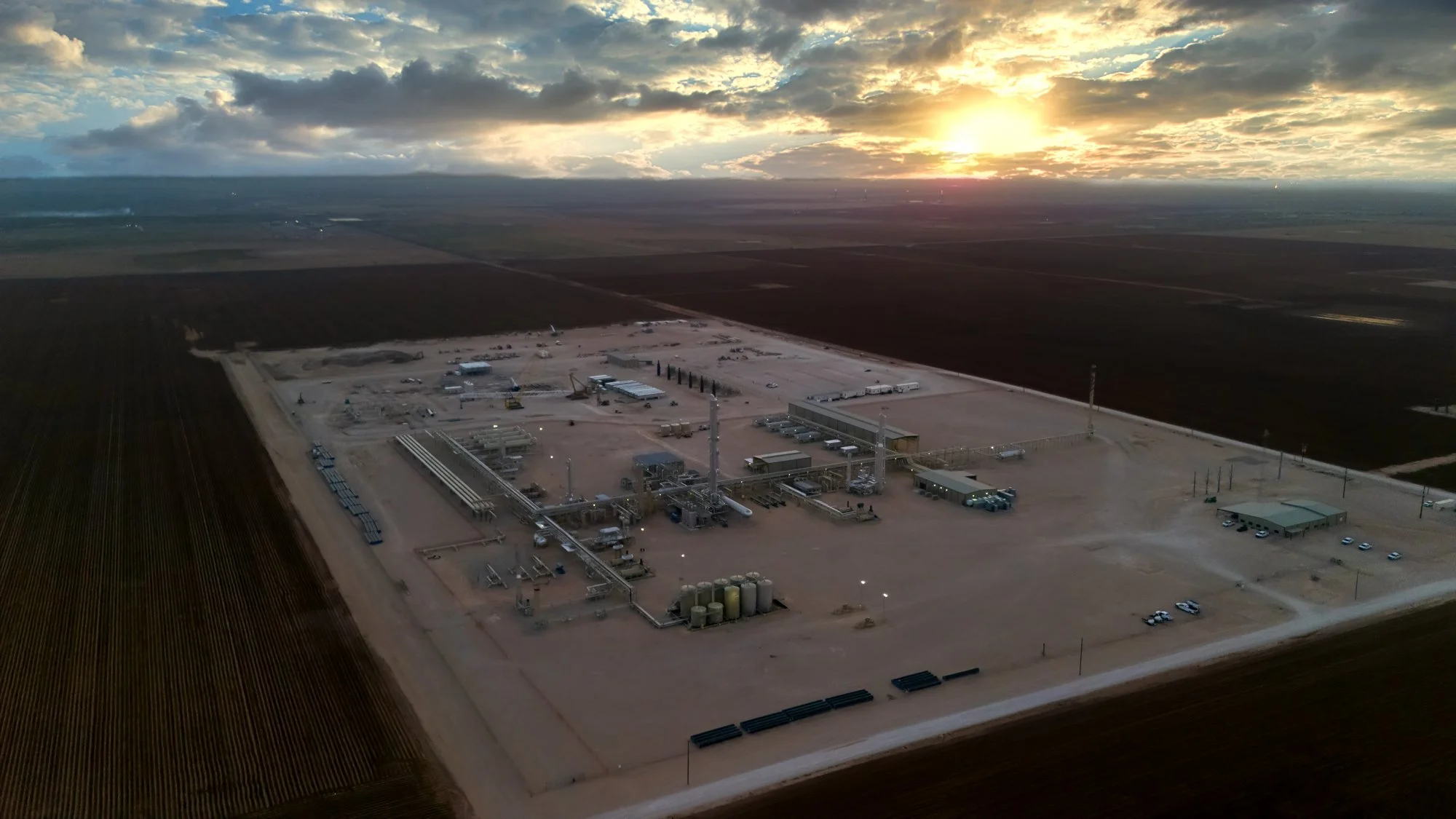 An aerial view of an industrial facility in a flat, rural area at sunset, surrounded by agricultural fields.
