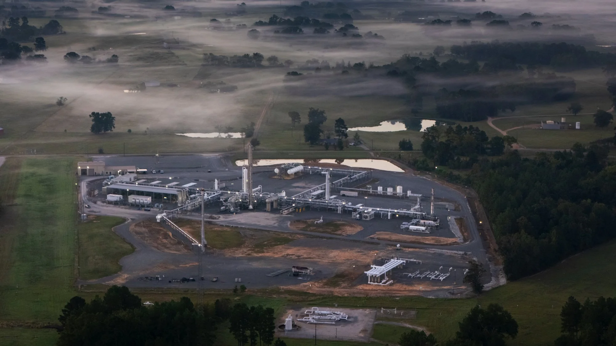 Aerial view of an industrial facility with pipelines, tall structures, and storage tanks, surrounded by green fields and trees, with fog in the background.