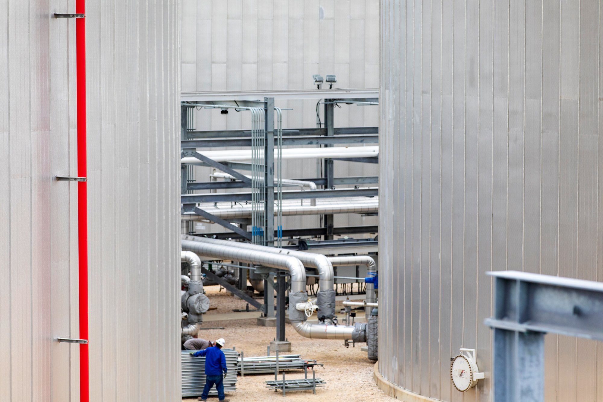 Industrial facility with large metal tanks and interconnected pipes, with two workers in hard hats and blue clothing working on construction.