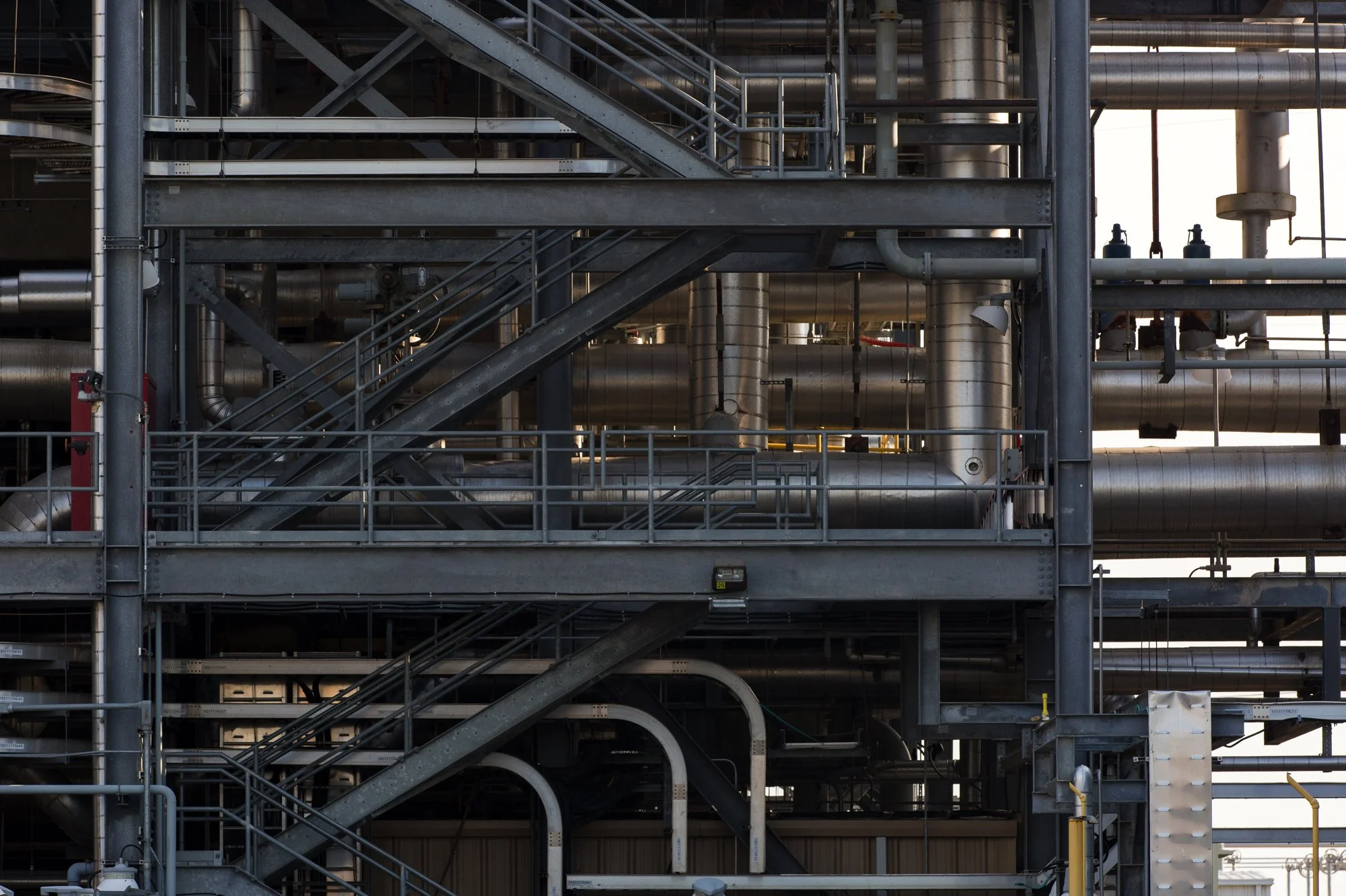 Industrial facility with multiple levels of metal walkways, stairs, and numerous large pipes running horizontally and vertically.