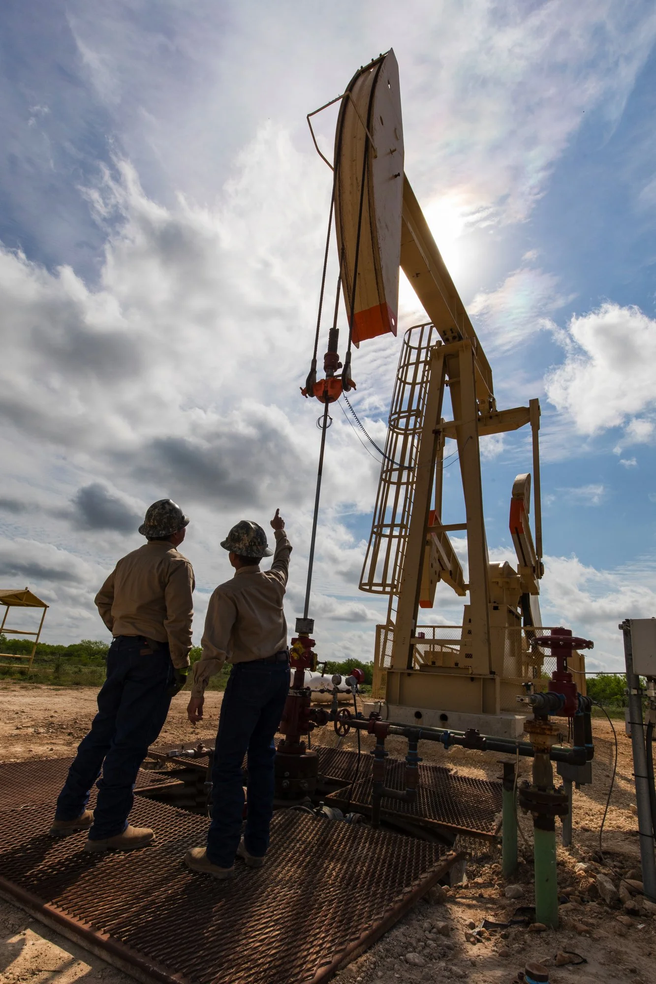 Two oil field workers in hard hats and work clothes stand near an oil pump jack in a field under a partly cloudy sky, engaged in discussion.