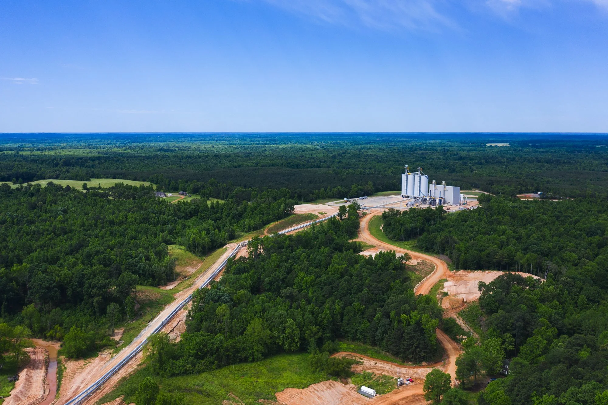 Aerial view of a rural area with dense forest, a winding dirt road, and industrial silos in the distance under a bright blue sky.