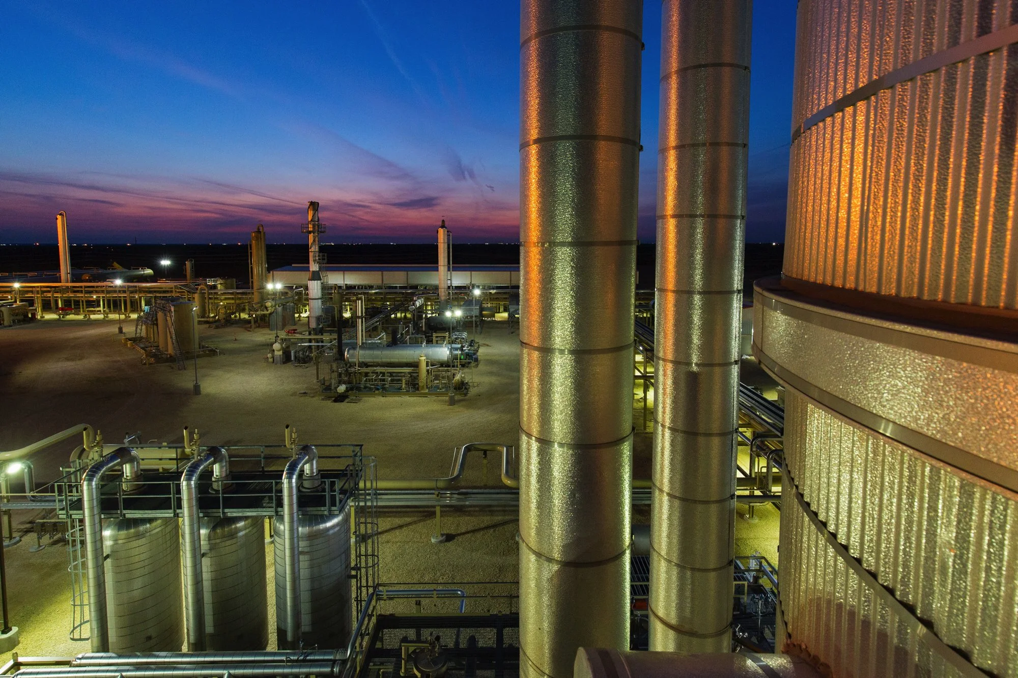 Industrial facility with large metallic pipes and tanks at sunset.