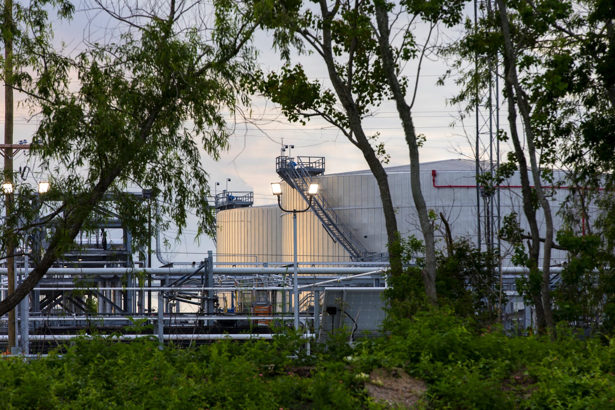 Industrial storage tanks and pipelines viewed through trees and greenery.