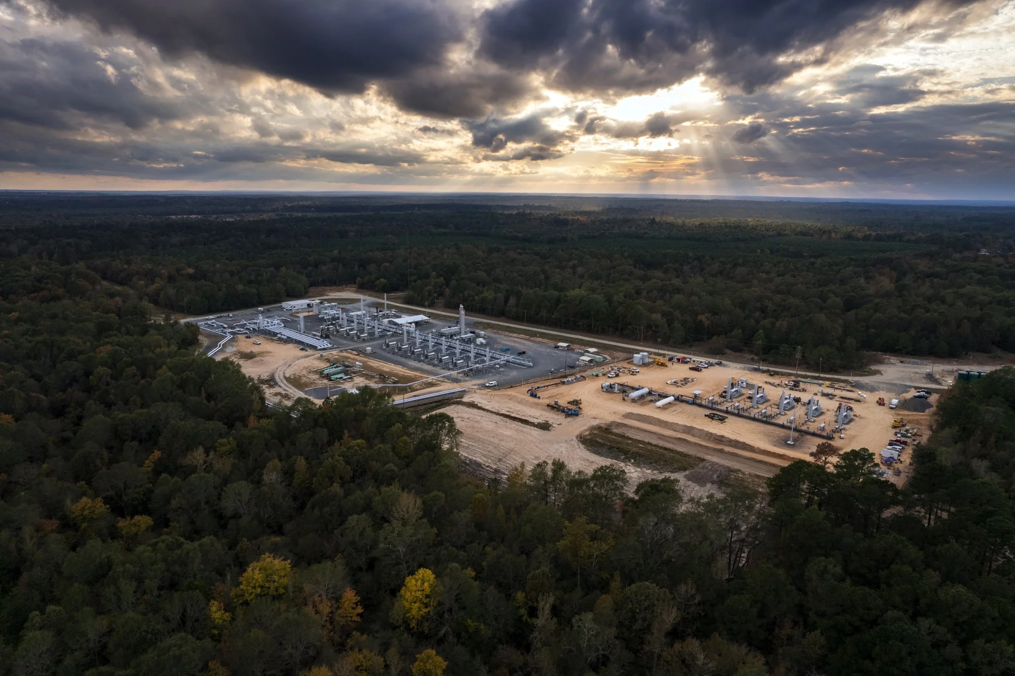An aerial view of an industrial facility surrounded by a forest, with cloudy skies and sunlight breaking through the clouds.