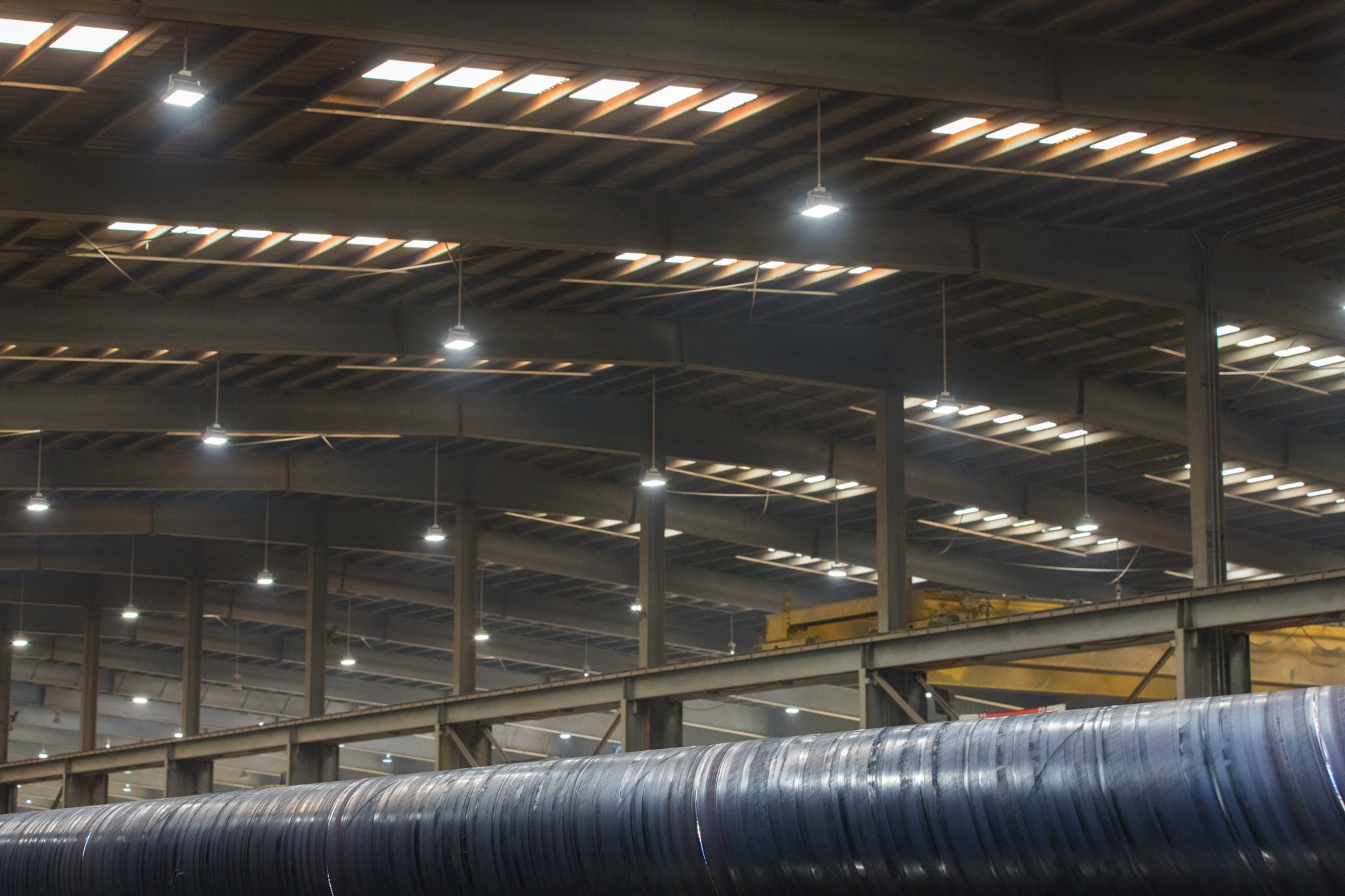 Inside a large industrial warehouse or factory with a high ceiling, exposed metal beams, and bright overhead lights. In the foreground, a large metallic pipe runs horizontally across the image.