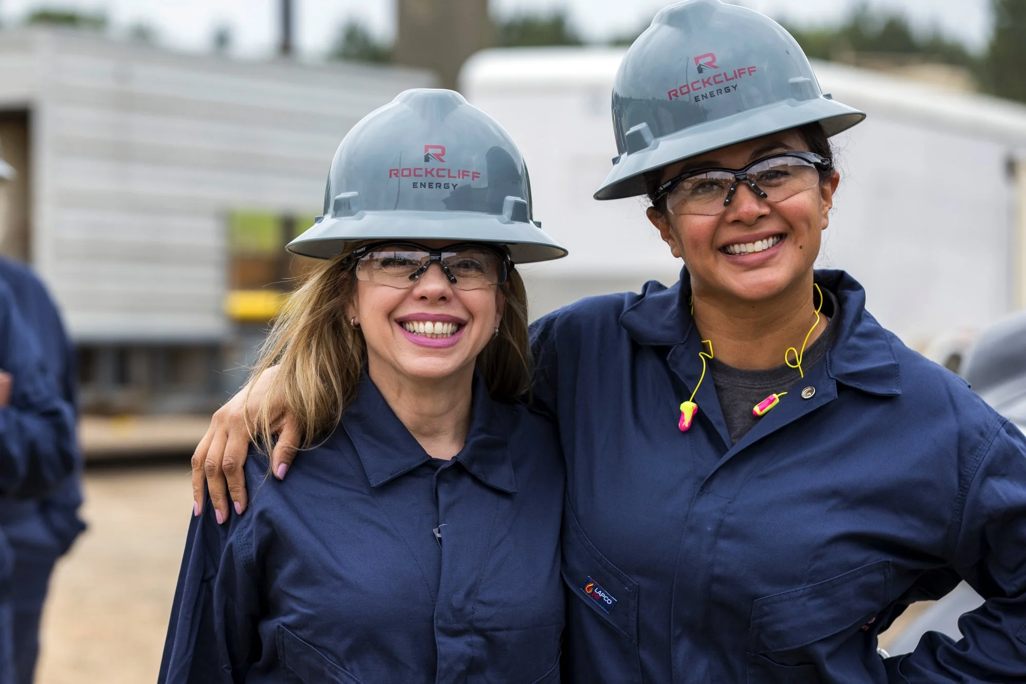 Two smiling women wearing helmets and safety glasses, standing outdoors at a construction site, with one woman's arm around the other.