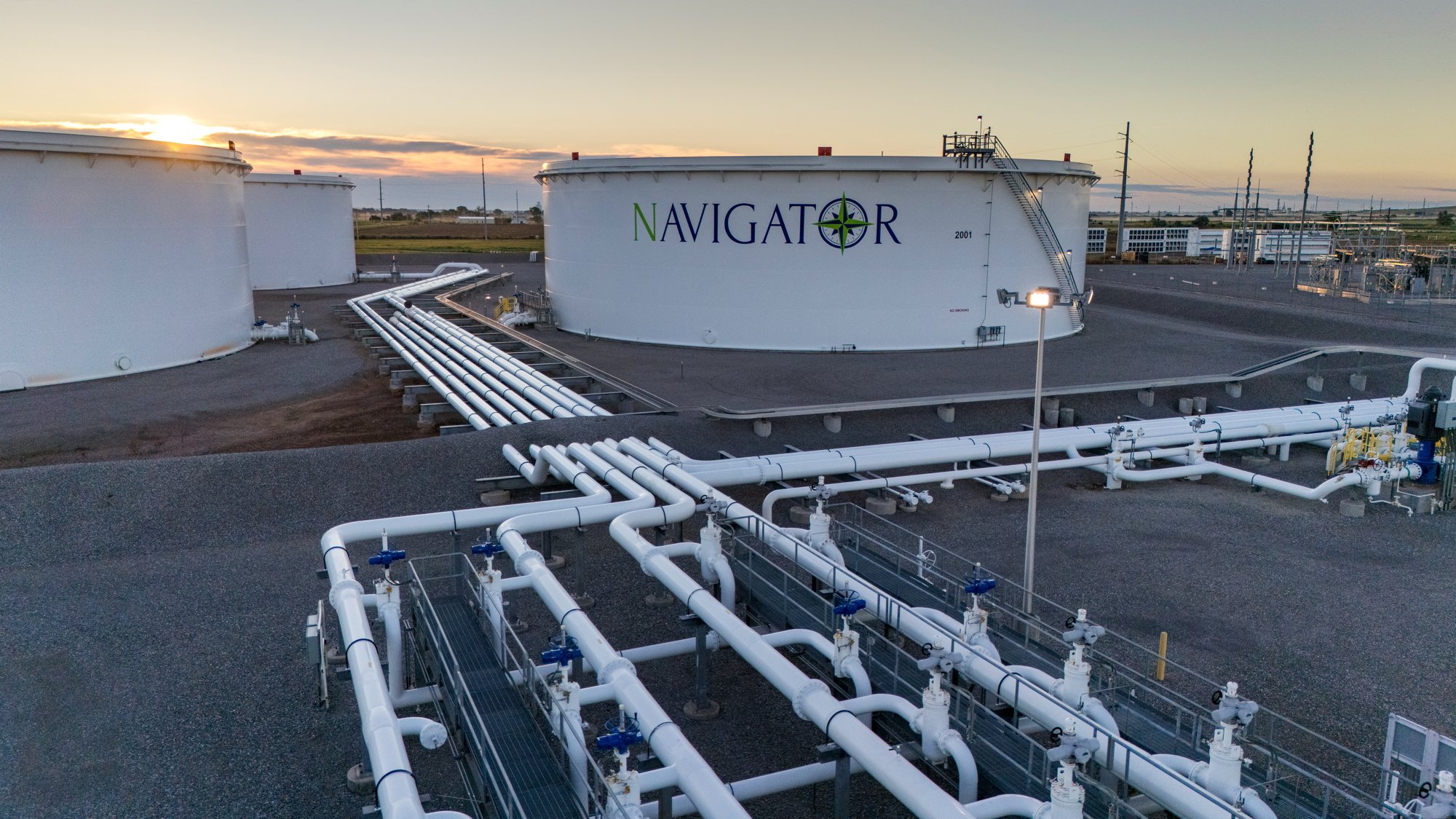 Large white fuel storage tanks with the label 'NAVIGATOR' in colorful letters and a green compass logo. Multiple white pipelines and valves run across the site, with some pipelines elevated and others at ground level. The surrounding area is an indus