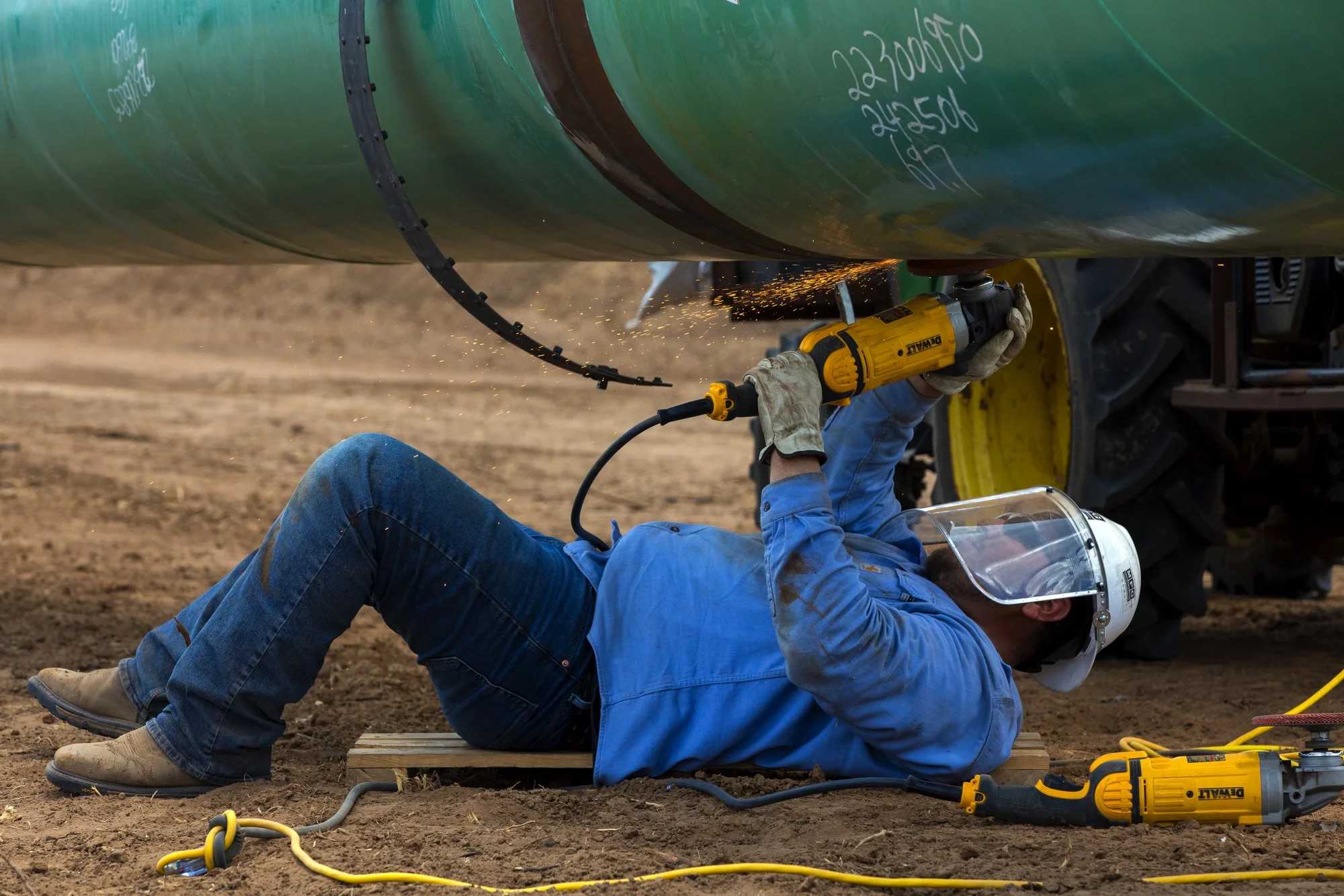 Worker lying underneath a large green pipeline, using a yellow power tool to cut or weld, sparks flying, on dirt ground with various cords around.