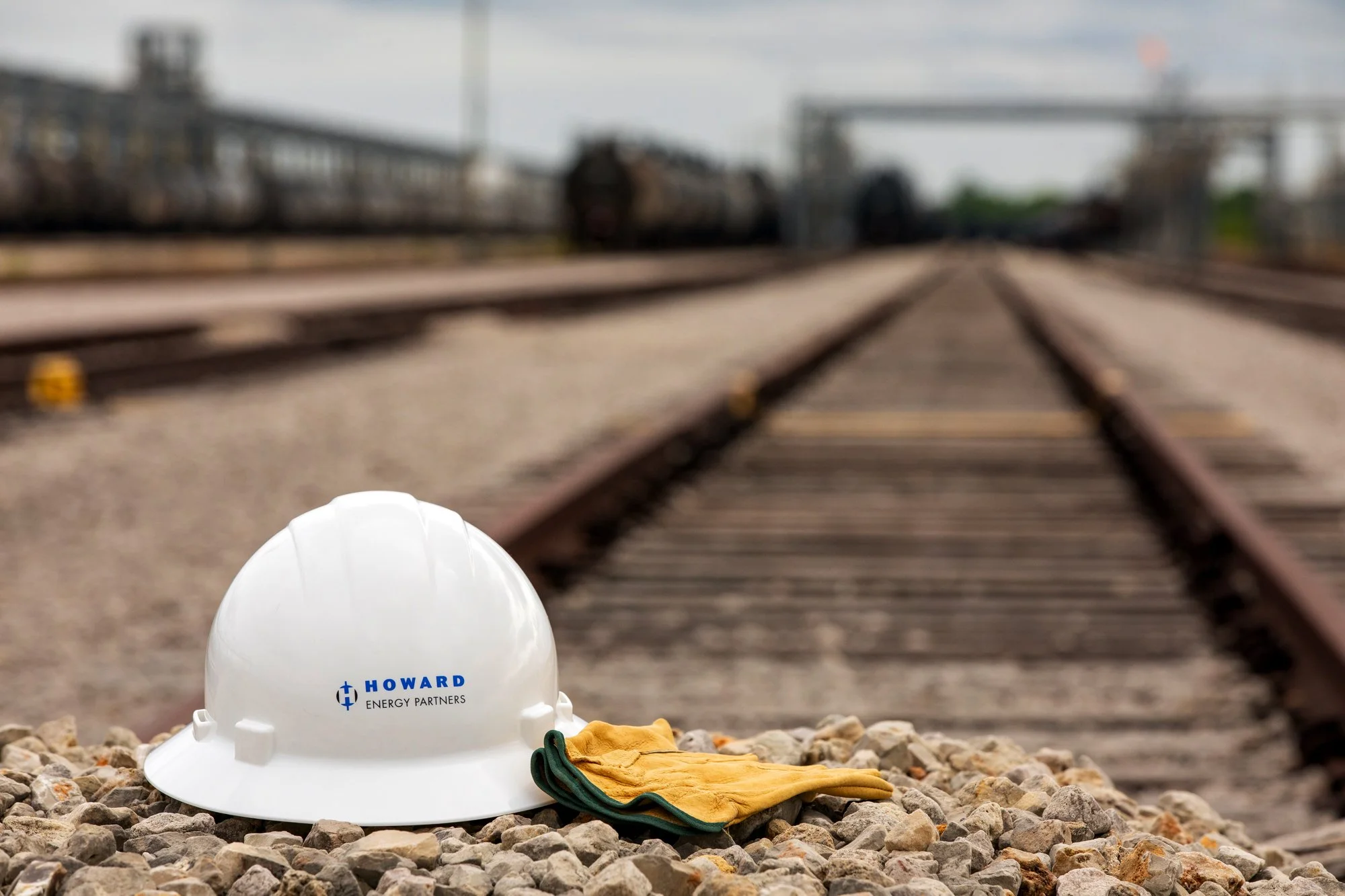 White safety helmet and yellow work gloves resting on gravel in front of railroad tracks with train cars in the distance.