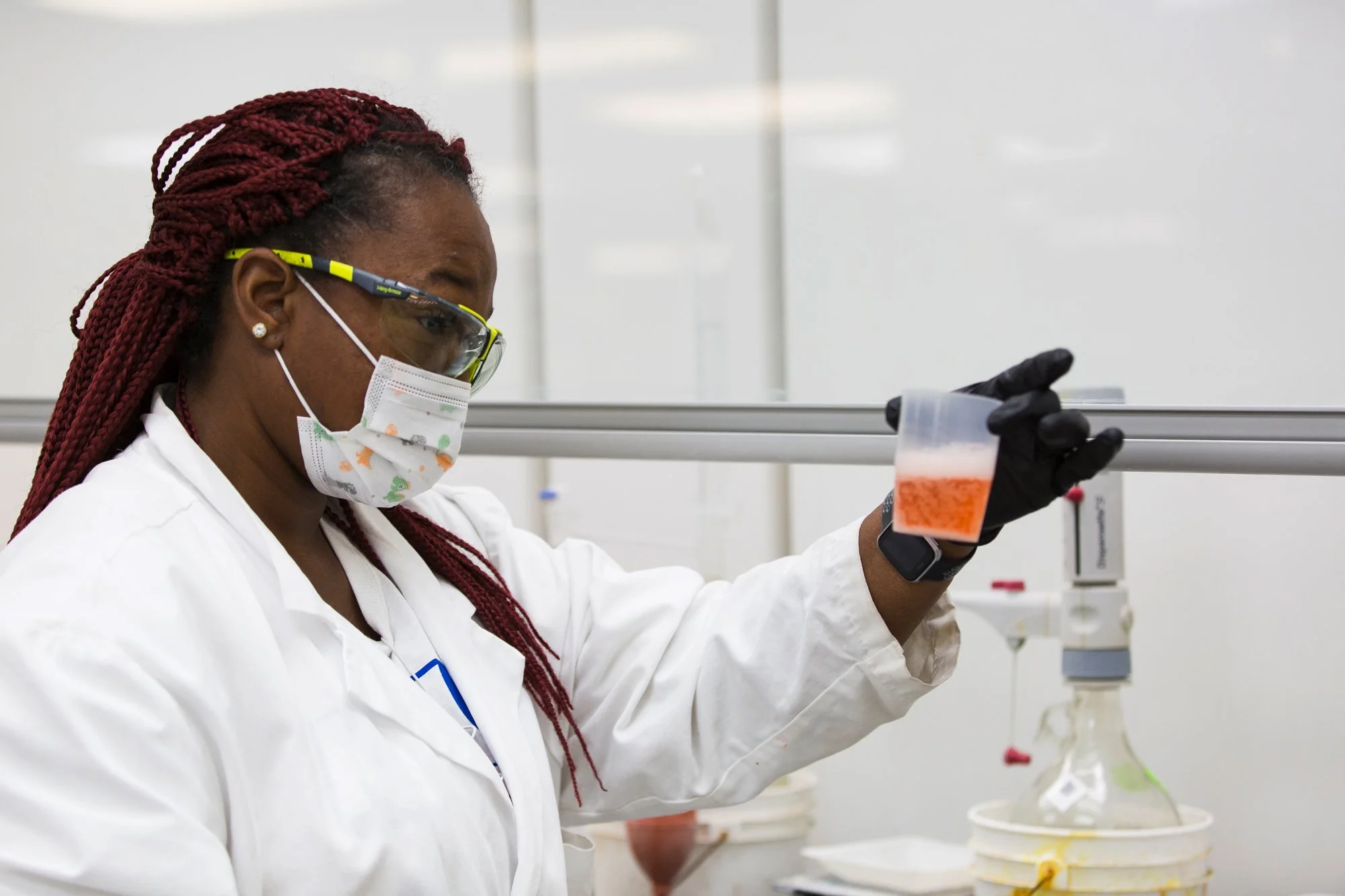 A scientist wearing safety goggles, a face mask, and gloves, holding a small container with orange substance in a laboratory.