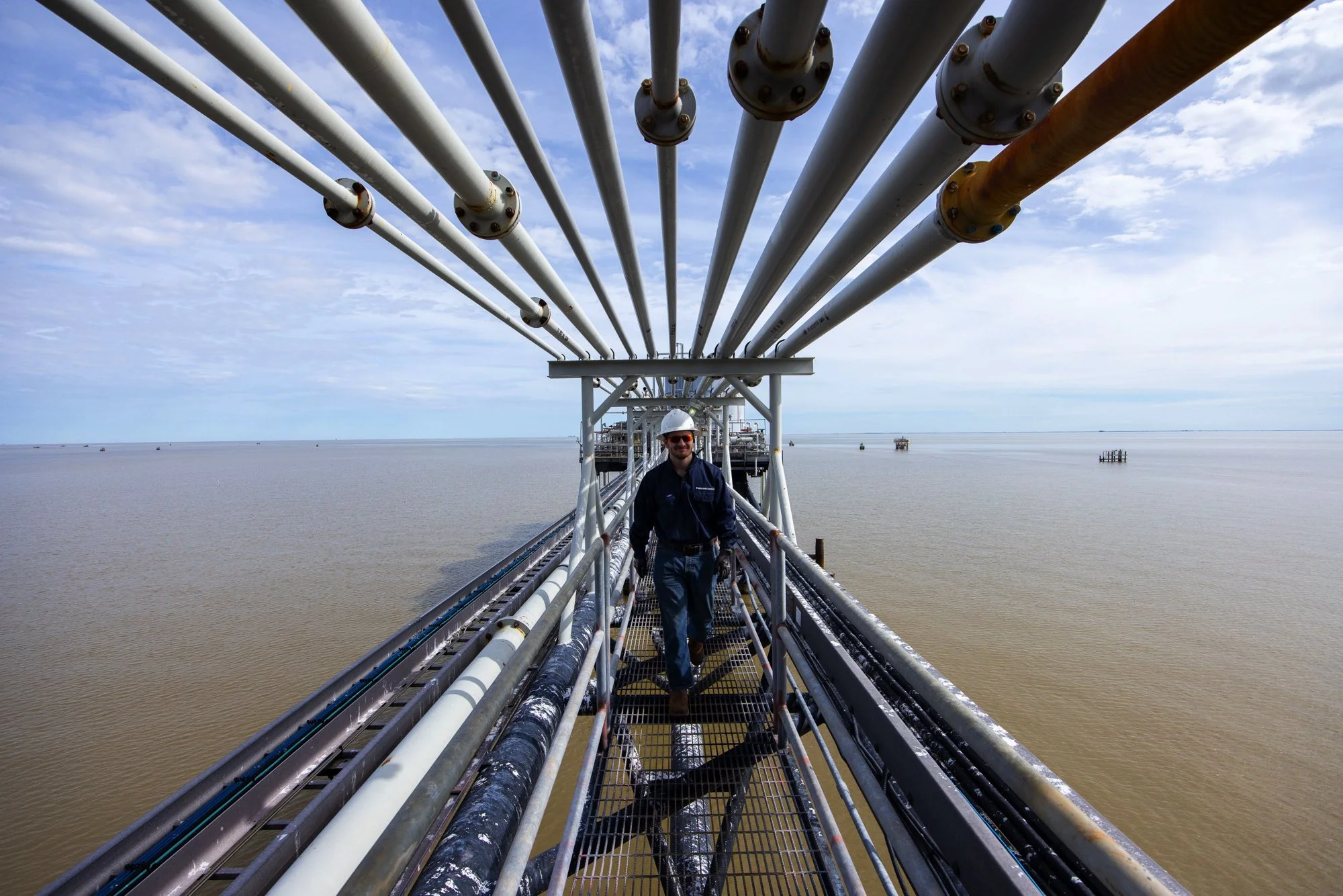A man wearing safety glasses, a helmet, and a dark jacket walking on a metal walkway on an oil rig or platform above the water with multiple pipelines overhead and a cloudy sky in the background.