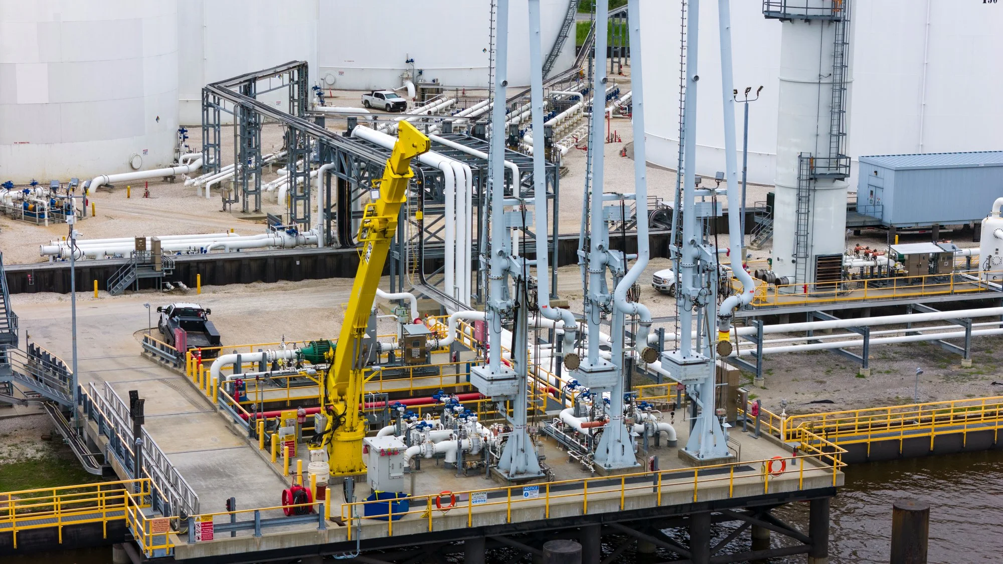 A large industrial facility by the water with numerous pipes, tanks, and a yellow robotic arm.