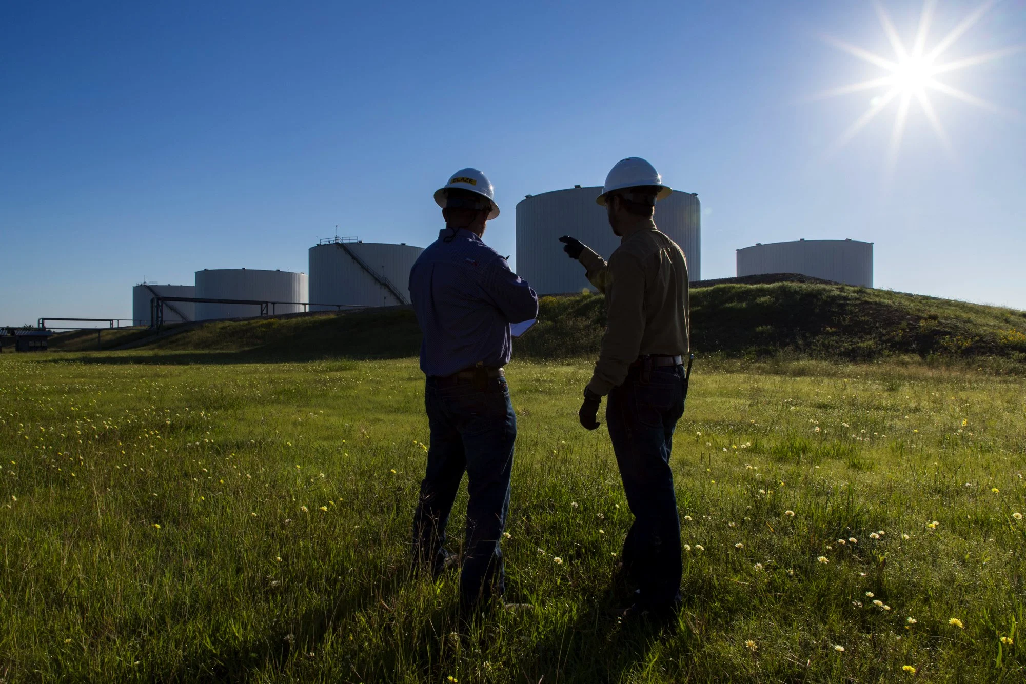 Two workers in hard hats and work clothes standing on a grassy field near large industrial storage tanks, with the sun shining brightly in the sky.