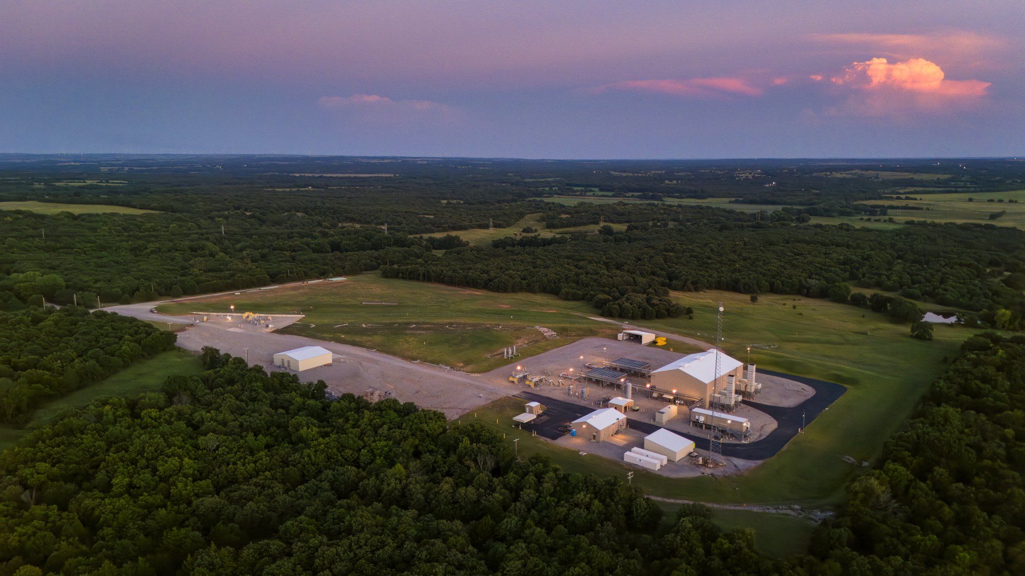 Aerial view of a large industrial facility in a rural area with dense forests and open fields, sunset lighting, and a cloudy sky.
