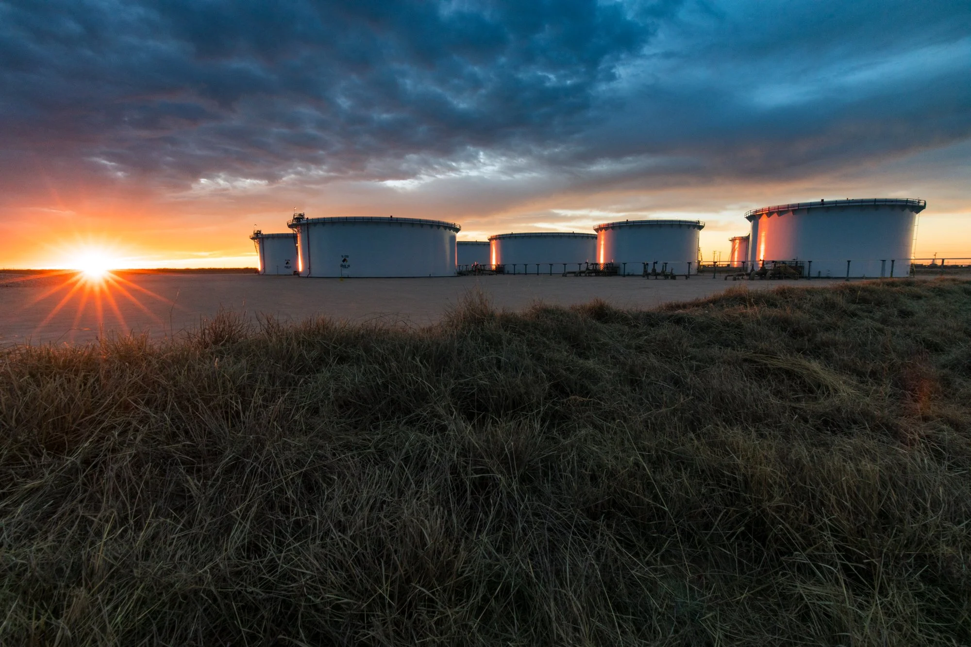 Six large white oil storage tanks in a flat open landscape with grassy foreground and cloudy sky at sunset.