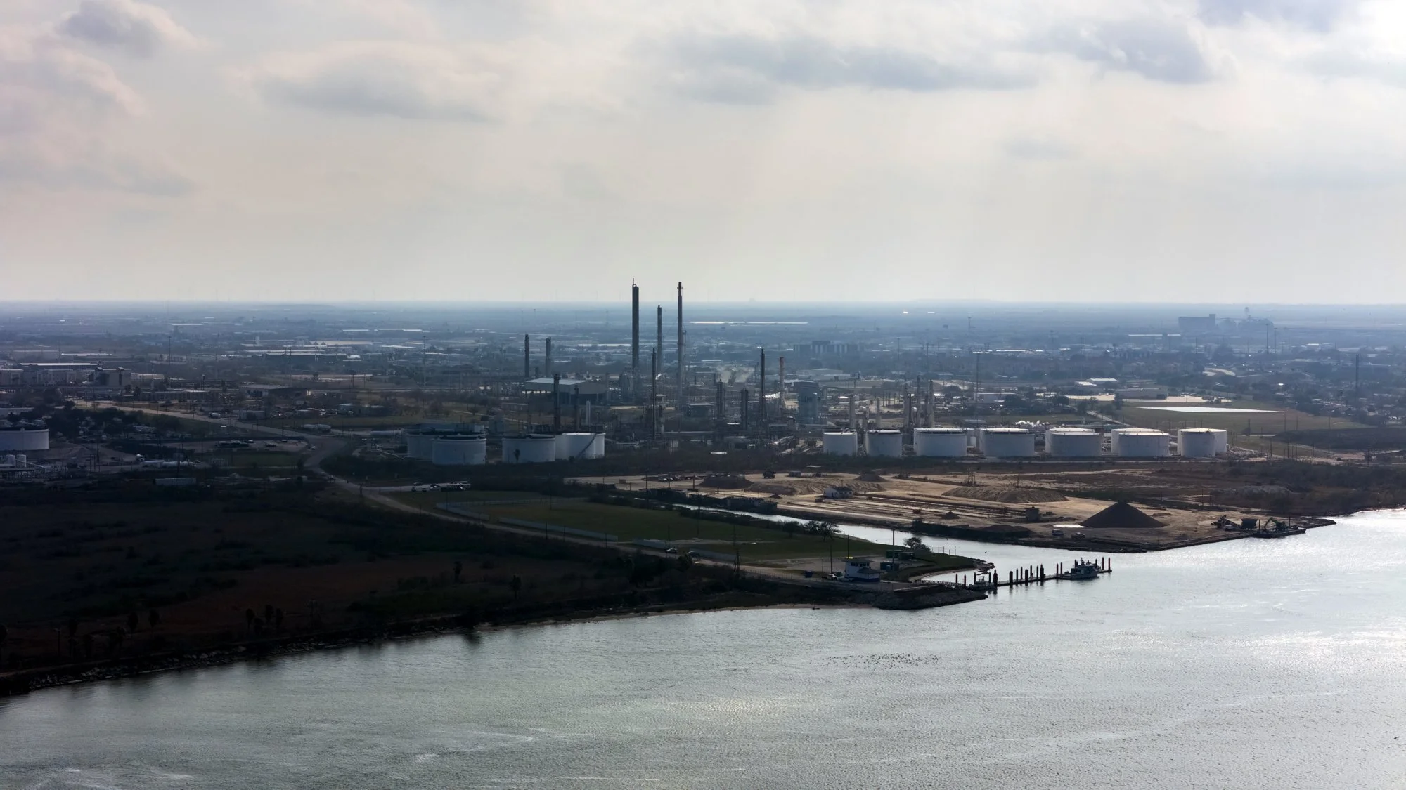 Aerial view of an industrial refinery near a body of water, with storage tanks, smokestacks, and infrastructure extending into the distance.