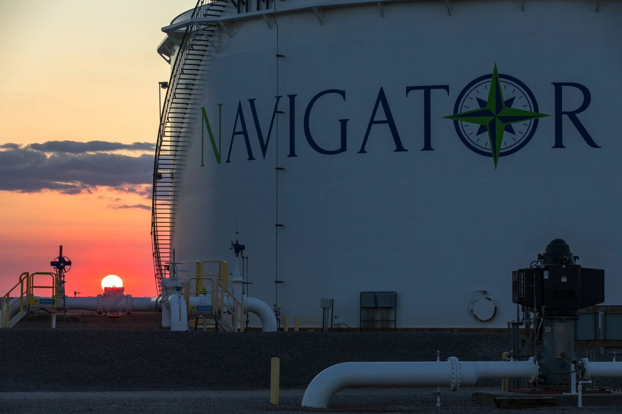 A large white industrial tank labeled 'Navigator' with a green compass logo, positioned outdoors at sunset or sunrise, with pipes and industrial equipment in the foreground.