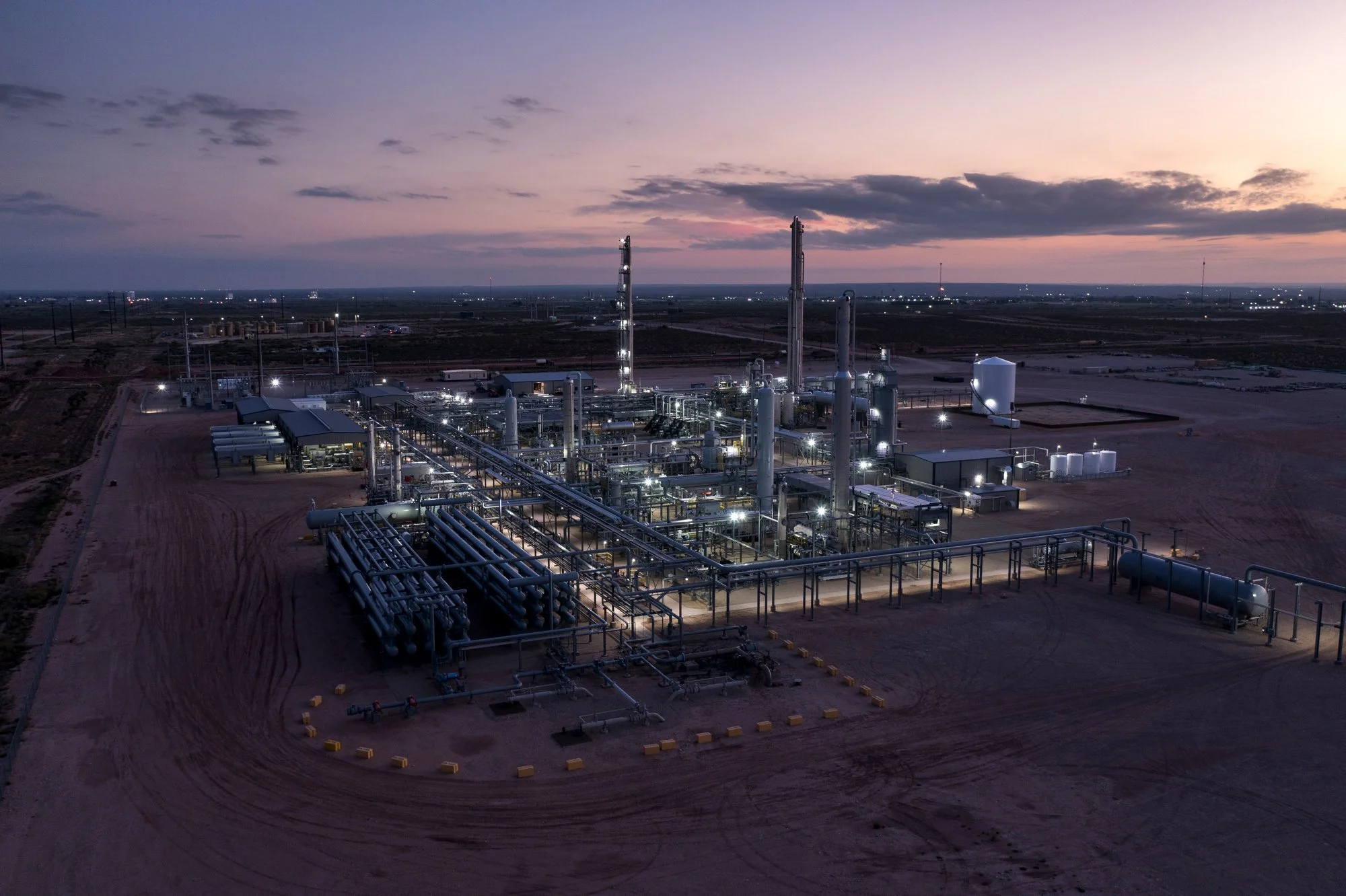 An aerial view of an industrial oil or gas processing facility at dusk with pipelines, tanks, and tall structures illuminated against a dusky sky.