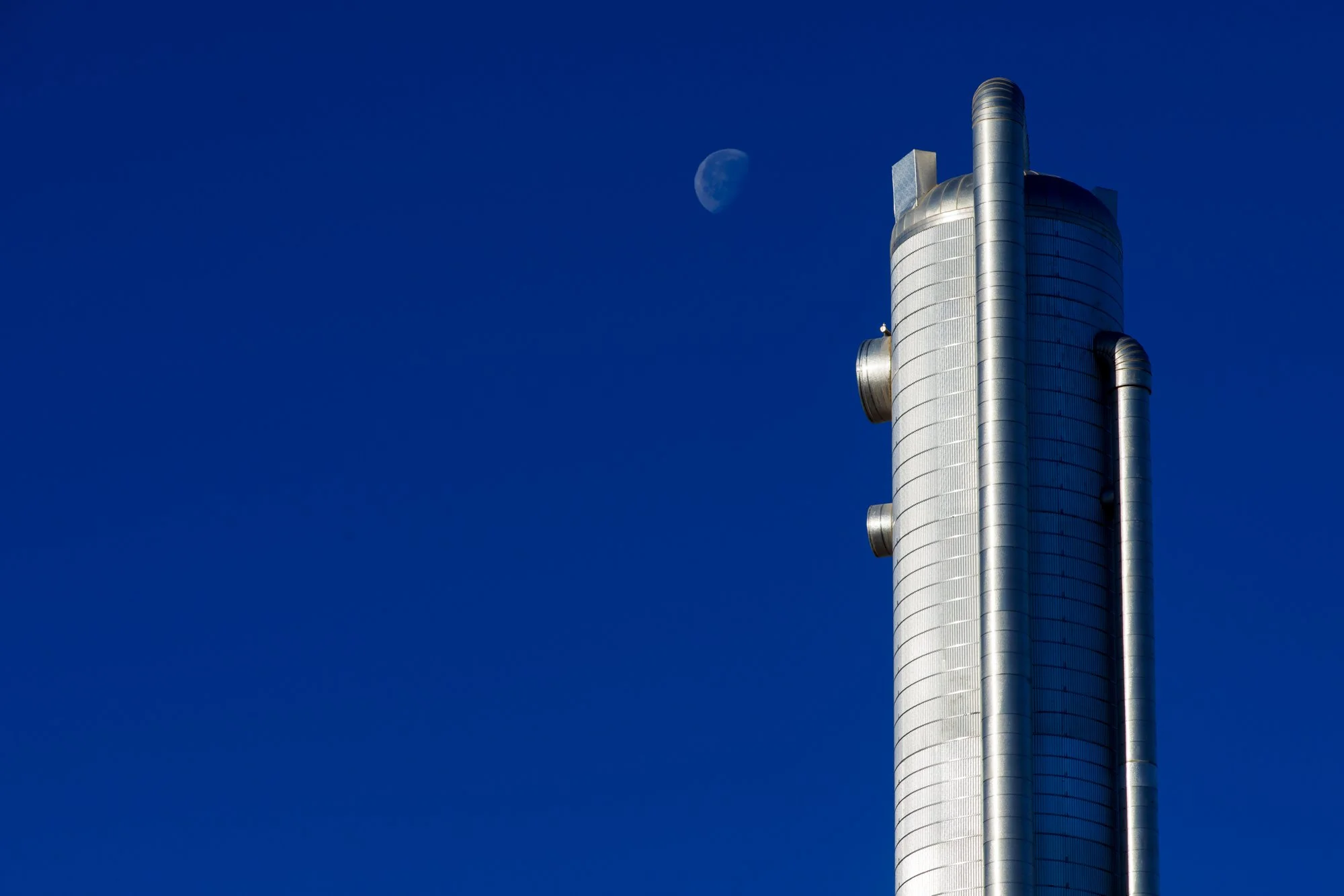 A tall, modern metallic building with rounded cylindrical shape and multiple protruding pipes against a bright blue sky with visible moon.