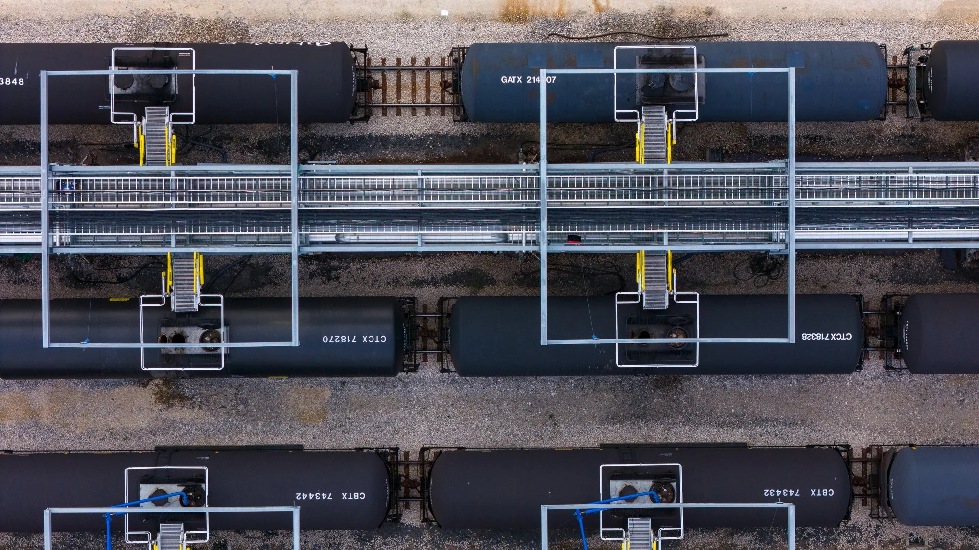 Top-down aerial view of a railroad yard with black tanker train cars parked on parallel tracks, some with visible identification numbers. White metal structures with stairs and walkways over the tracks are present, positioned above the cars. The grou
