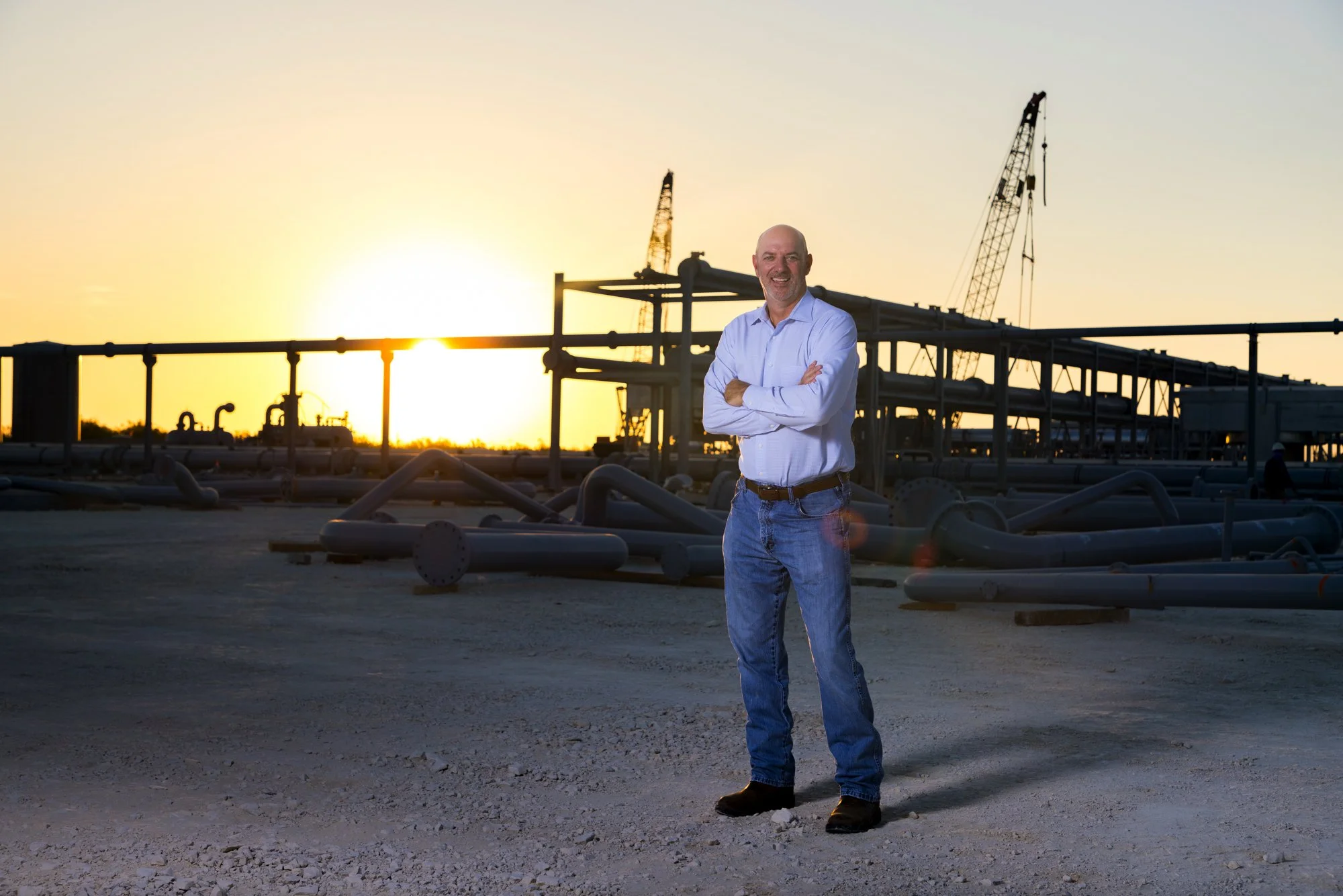 A man in a white shirt and jeans smiling with arms crossed at an oil drilling site during sunset.