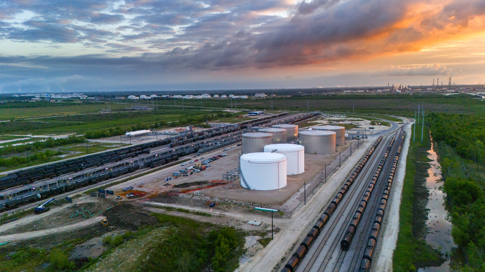 An aerial view of a large industrial area with storage tanks, oil refinery structures, railroad tracks with freight trains, and surrounding green fields and water bodies during sunset.