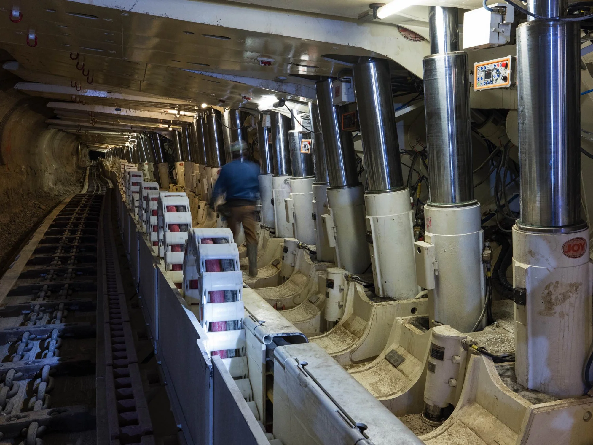 A worker in blue shirt and beige pants walking along side tunnel boring machine with large steel and white components, inside a tunnel with track and soil on the sides.