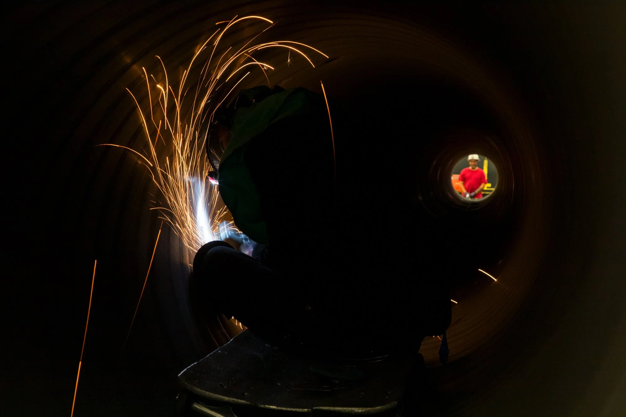 A worker welding inside a large pipe, with sparks flying, and a person visible through the pipe's opening in the background.