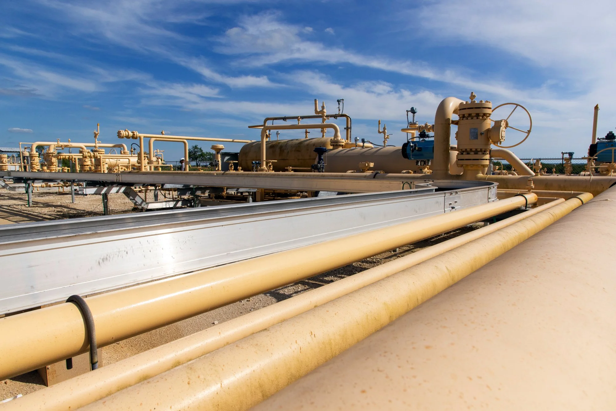 Aerial view of industrial pipelines and equipment in an oil or gas processing facility under a blue sky.