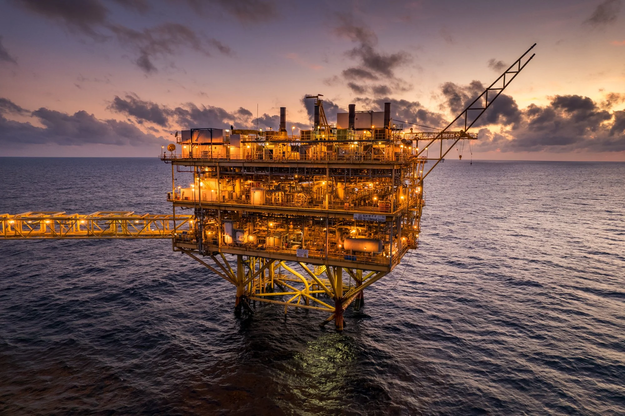 Offshore oil rig illuminated at sunset in the ocean with clouds in the sky.