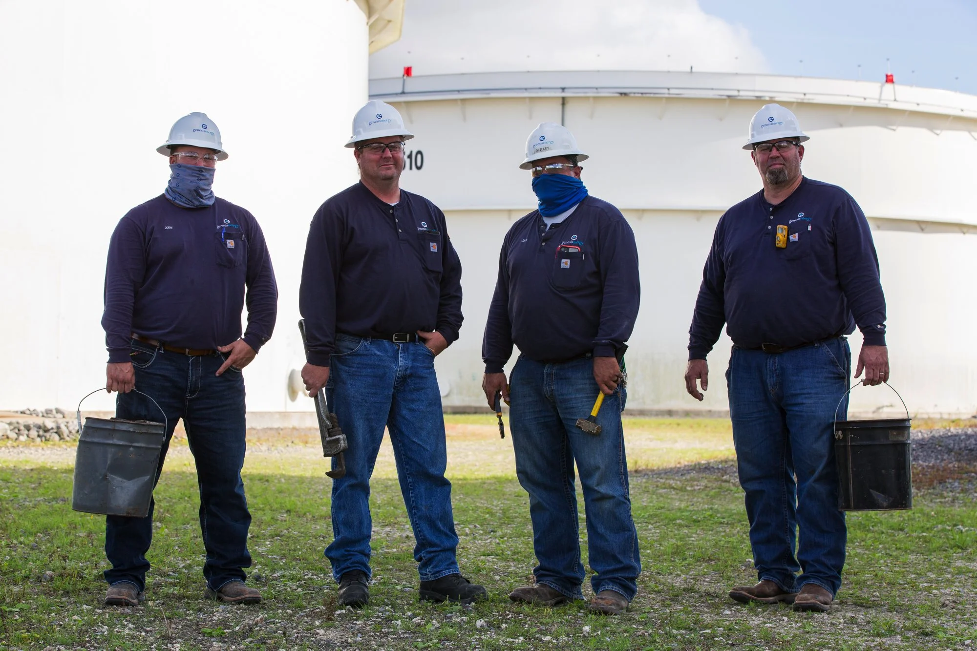 Four workers standing outdoors in front of a large industrial storage tank, wearing helmets, safety glasses, and blue uniforms, with tools and buckets.