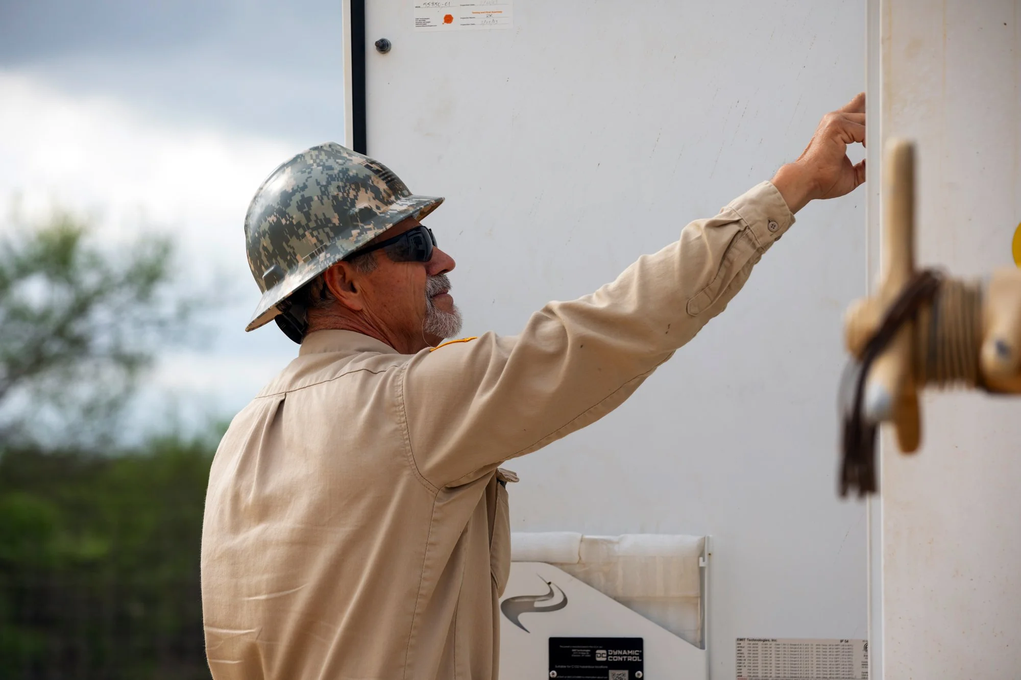 A man in military-style camouflage helmet and sunglasses reaching for something on a whiteboard outdoors.