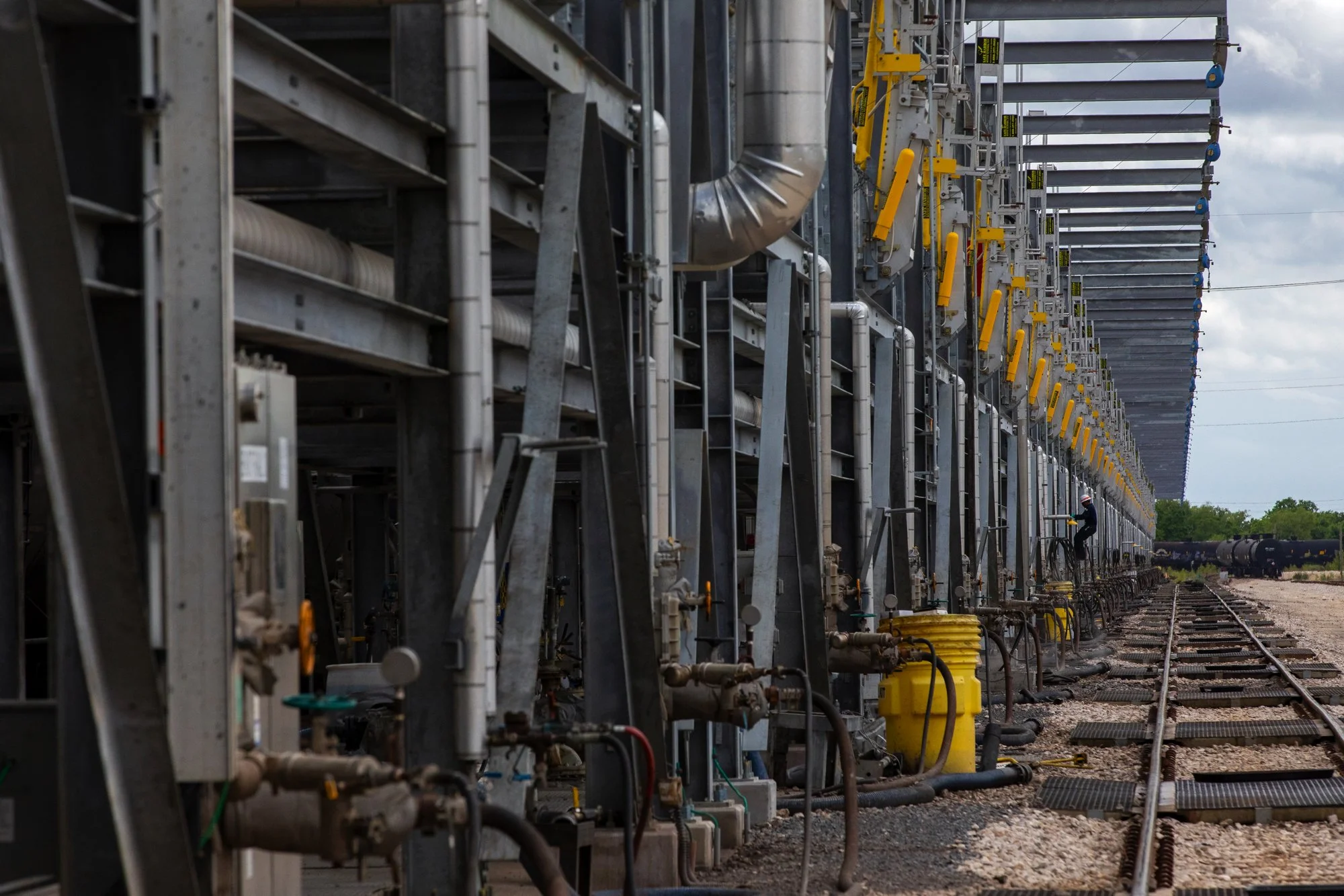 Industrial rail yard with metal pipes, valves, and equipment along the tracks under a cloudy sky, with a worker in safety gear inspecting the setup.