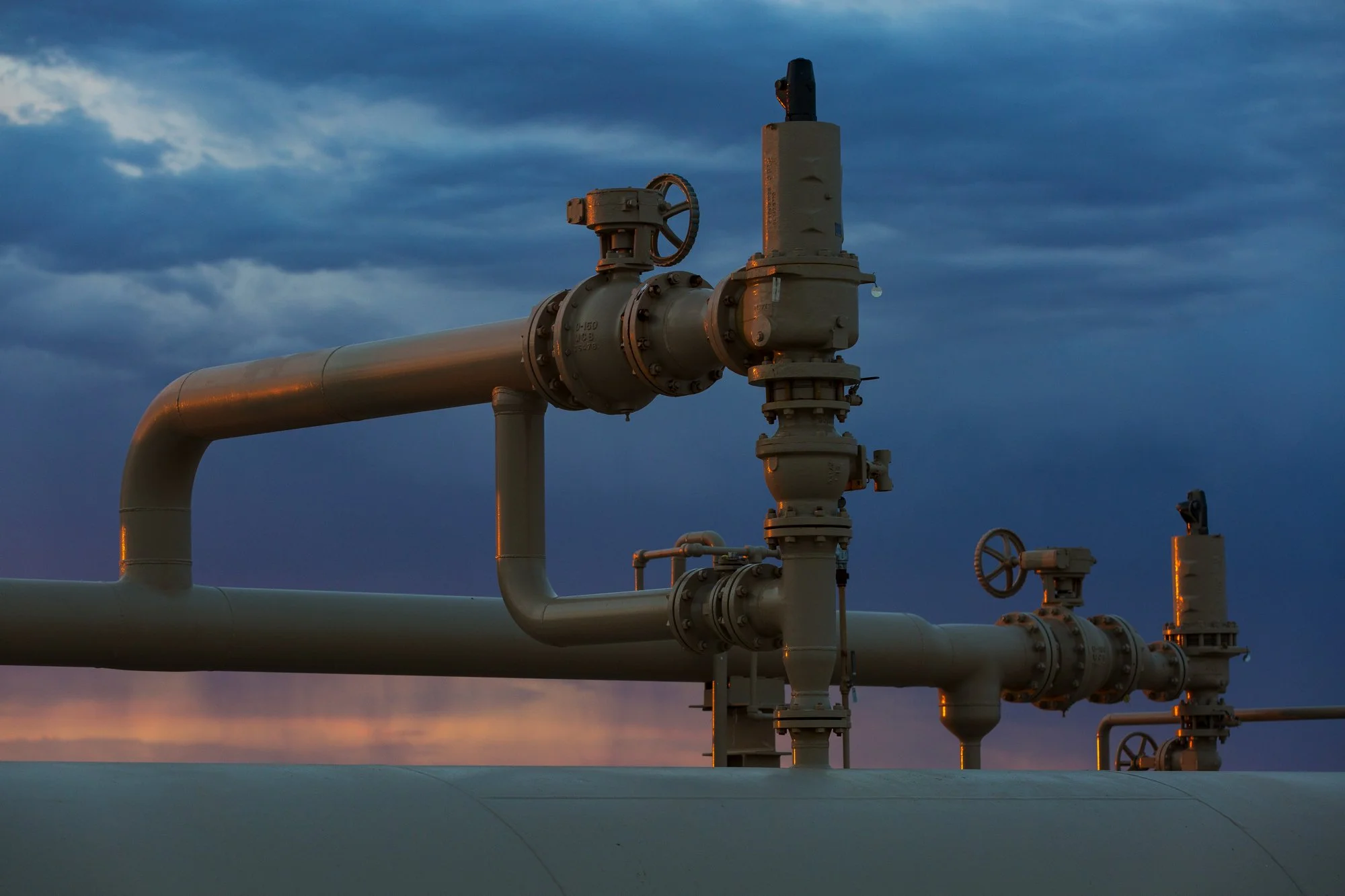 Close-up of industrial pipelines with valves against a cloudy sky at dusk.