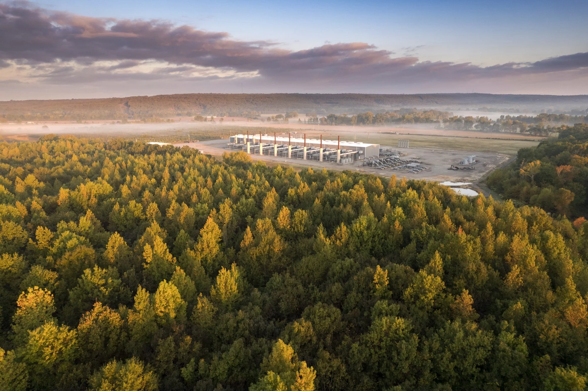 An aerial view of a power plant with multiple smokestacks surrounded by a dense green forest, with mist and clouds in the sky.