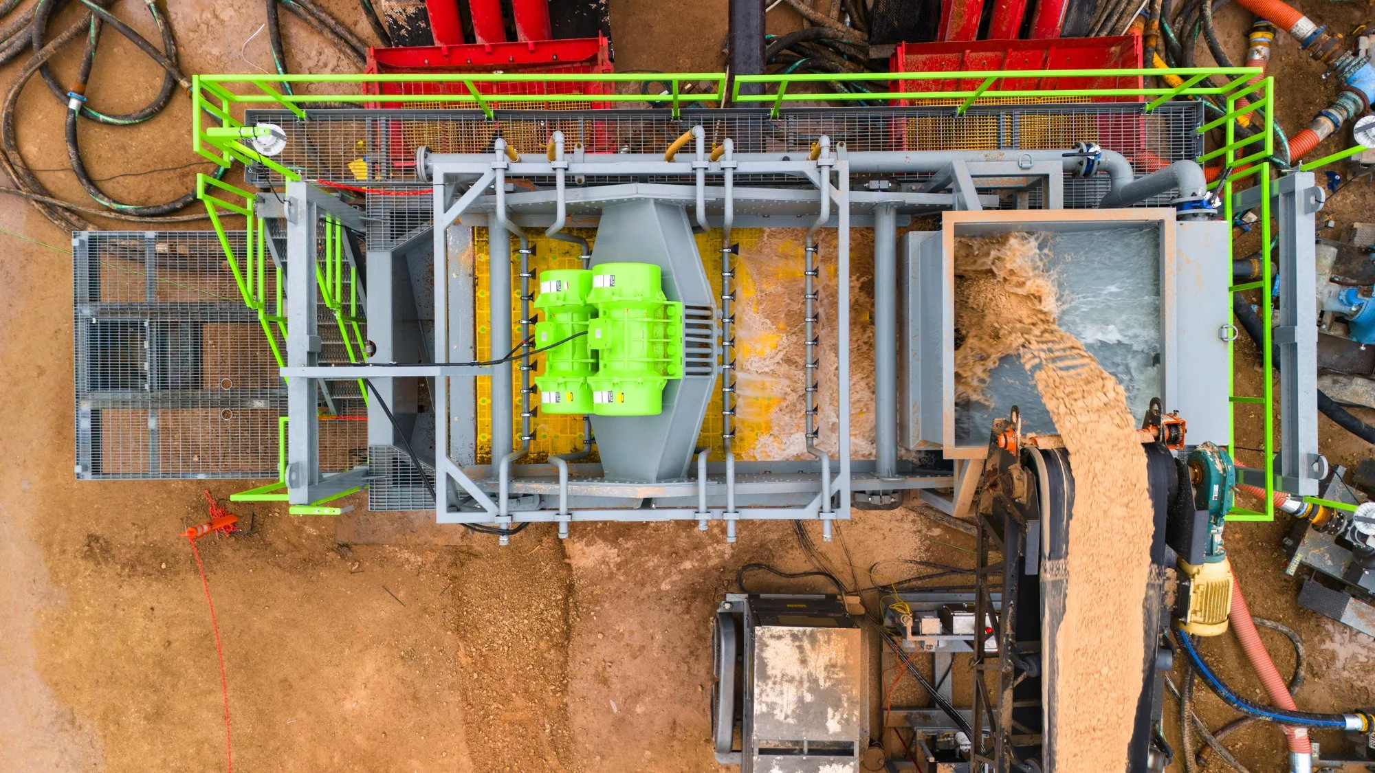 Overhead view of a construction site showing a large piece of industrial machinery pouring a stream of sand into a container, with green safety railings and surrounding pipes and equipment.