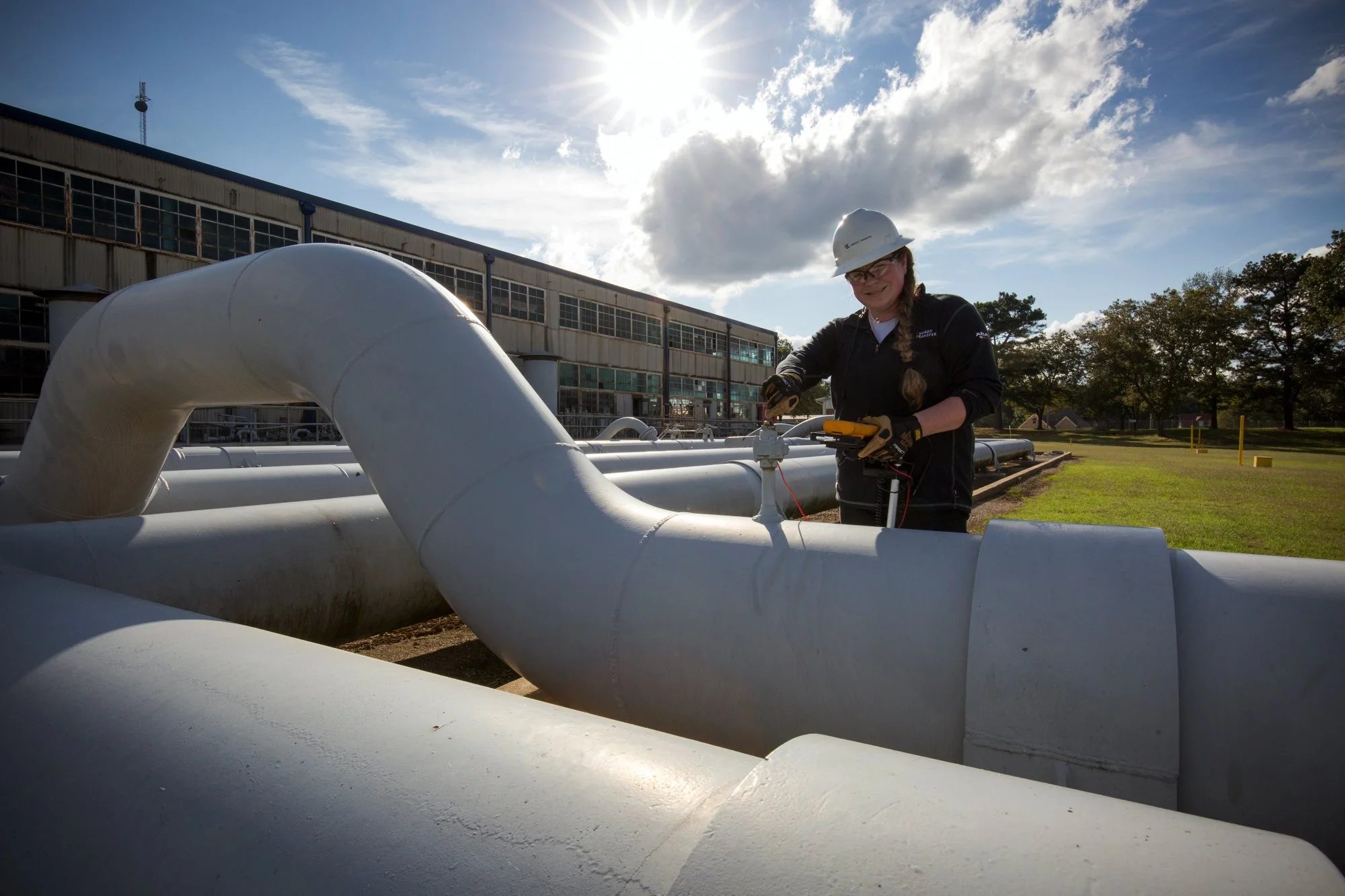 A woman wearing a white hard hat and safety gloves is working on a large white industrial pipe outside of a facility on a sunny day.