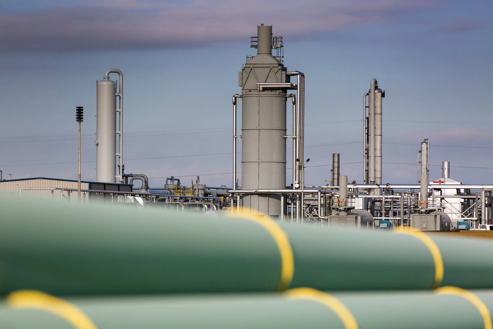 Industrial refinery with large vertical tanks and pipes under a cloudy sky, foreground includes blurred green pipeline with yellow markings.